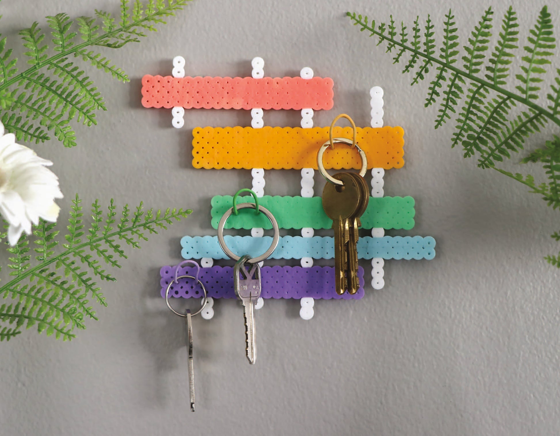 The Melt and Make Bead Crafts Digital Download by Leisure Arts features a colorful key holder, displayed on a gray wall holding three sets of keys, with green ferns and a white flower—great inspiration for beginner projects.