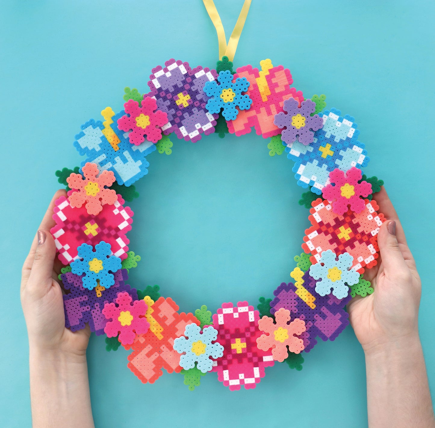 Two hands hold a vibrant floral wreath made with Melt and Make Bead Crafts Digital Download by Leisure Arts, featuring pink, purple, blue, and yellow flowers on turquoise. A yellow ribbon tops this fun beginner craft project.