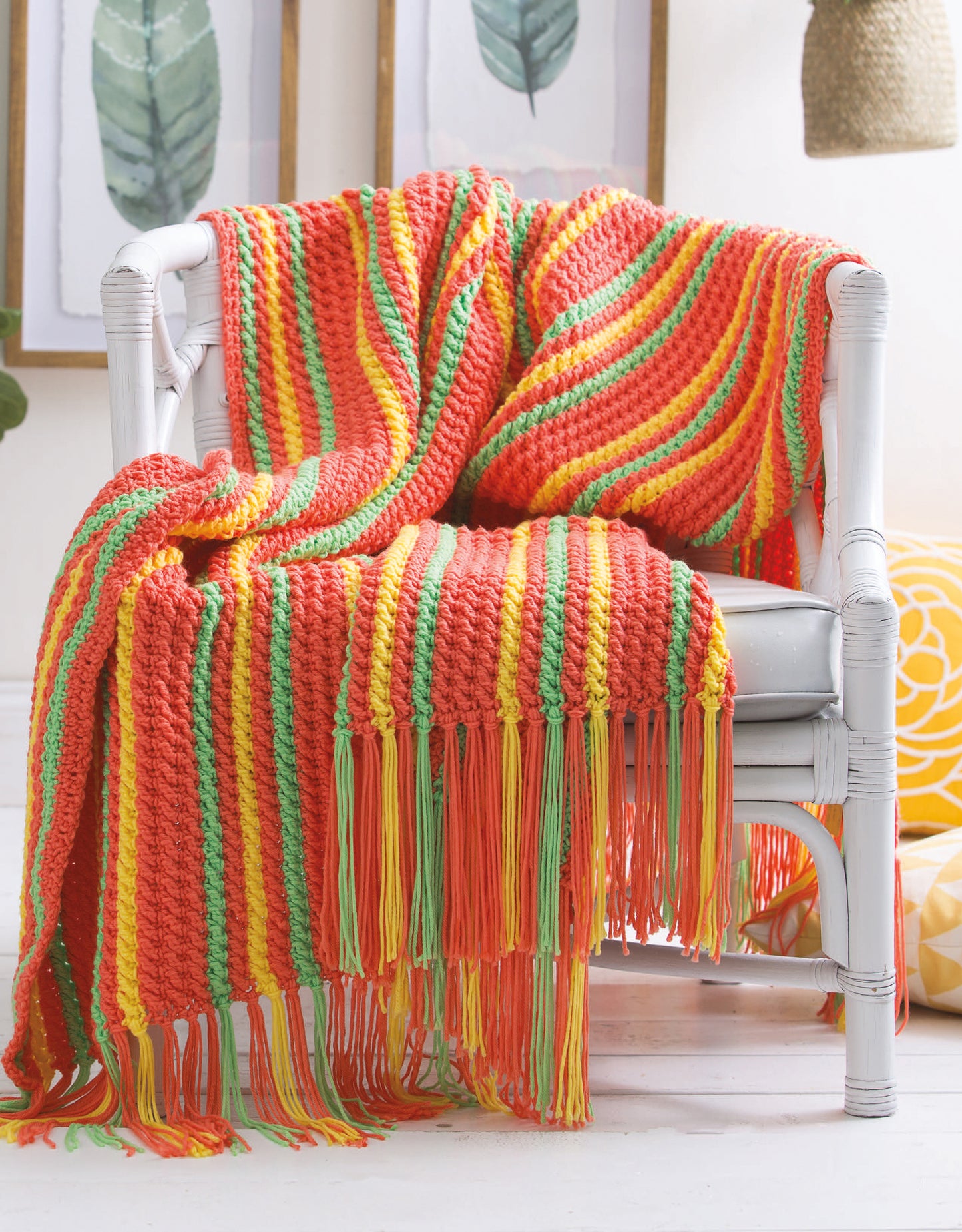 The Leisure Arts Bright Afghans Crochet blanket, featuring orange, yellow, and green stripes with long fringe, is draped over a white chair in a sunlit room with framed botanical prints on the wall.