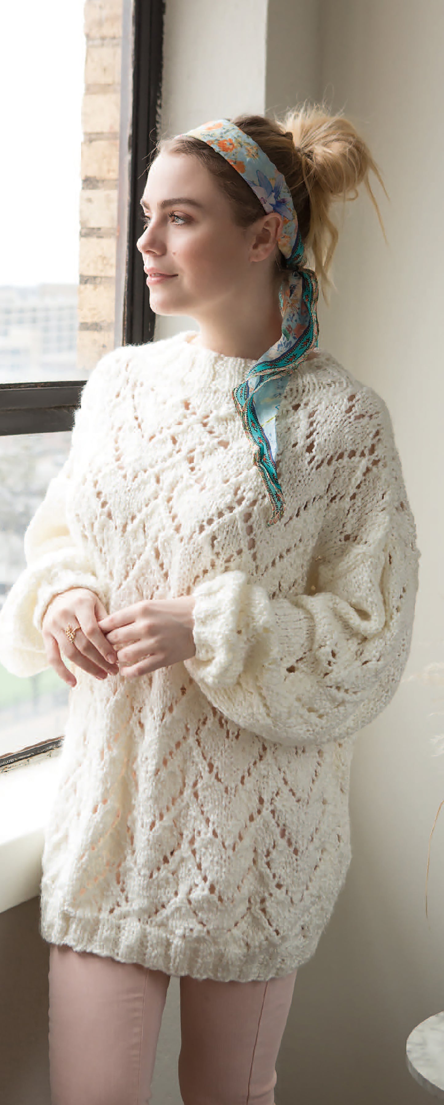 A woman stands by a window in calm reflection, wearing the Leisure Arts Sweater Weather Knit's cream chunky sweater and a colorful scarf tied in her hair, hands clasped gently in front of her.