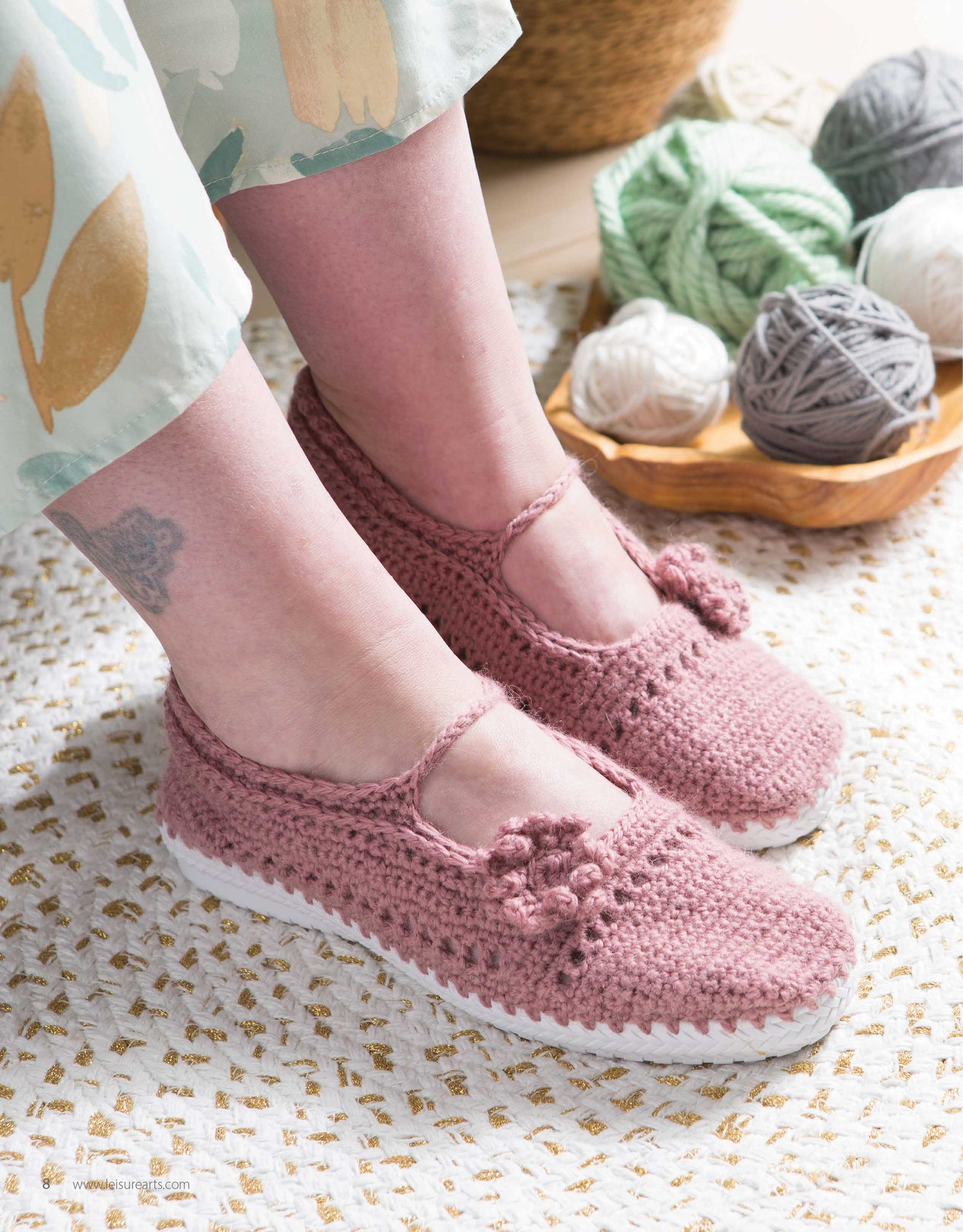 Close-up of feet in pink Crochet Shoes by Leisure Arts, featuring a small flower detail, standing on a woven mat. Balls of yarn and crochet tools appear in the background.