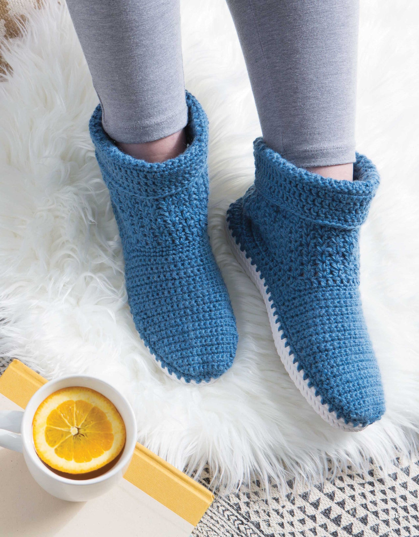 A person in gray leggings relaxes on a white rug, wearing Leisure Arts Crochet Shoes—blue crocheted slipper boots—beside a mug with lemon water and a partially visible yellow book.