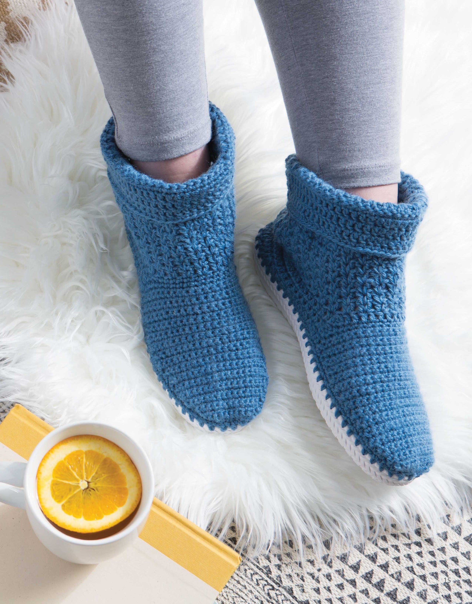 A person in gray leggings relaxes on a white rug, wearing Leisure Arts Crochet Shoes—blue crocheted slipper boots—beside a mug with lemon water and a partially visible yellow book.