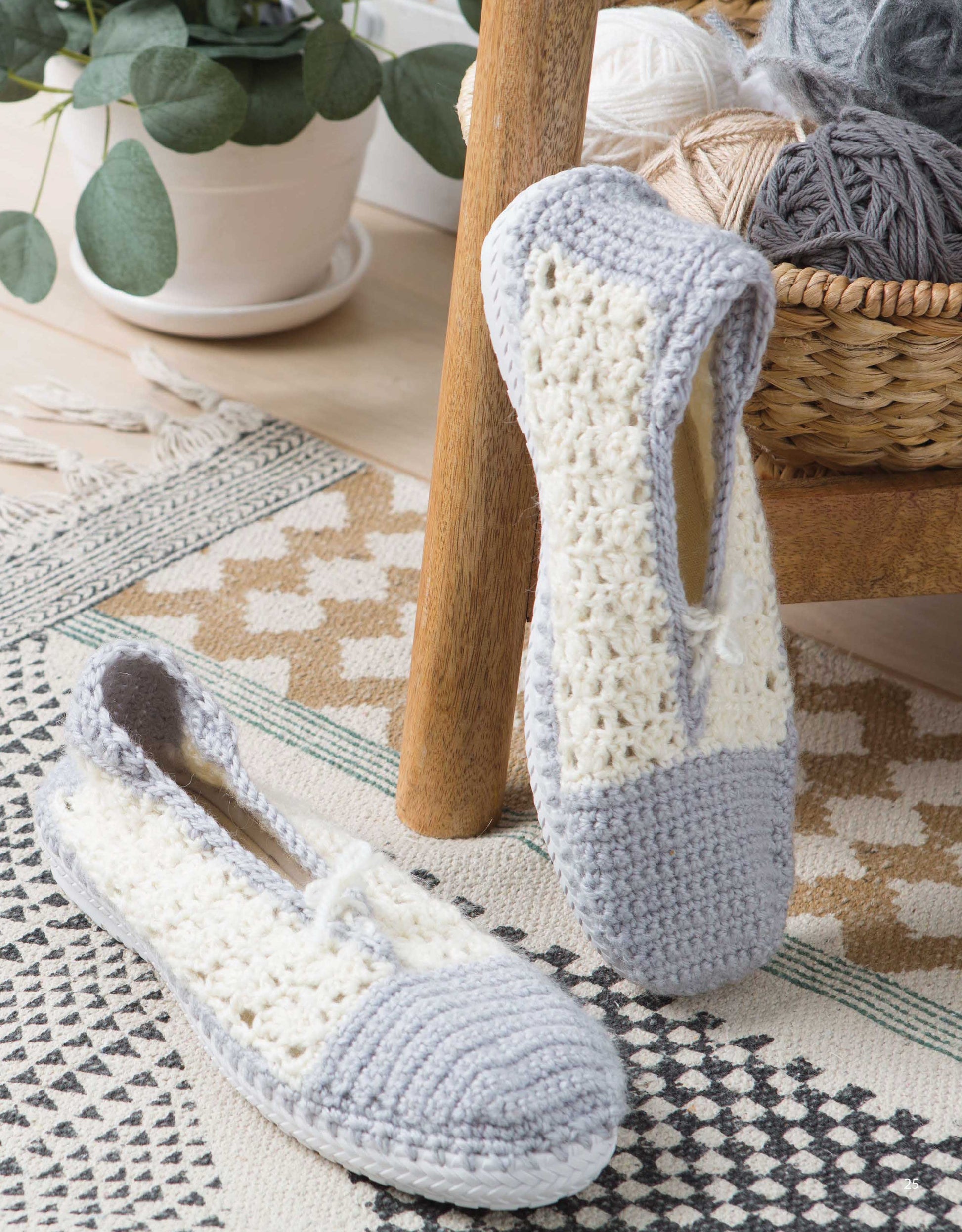 Leisure Arts Crochet Shoes featuring gray toes and heels with cream sides rest on a patterned rug by a wooden chair—cozy, stylish footwear. A basket of yarn and a potted plant are visible in the background.