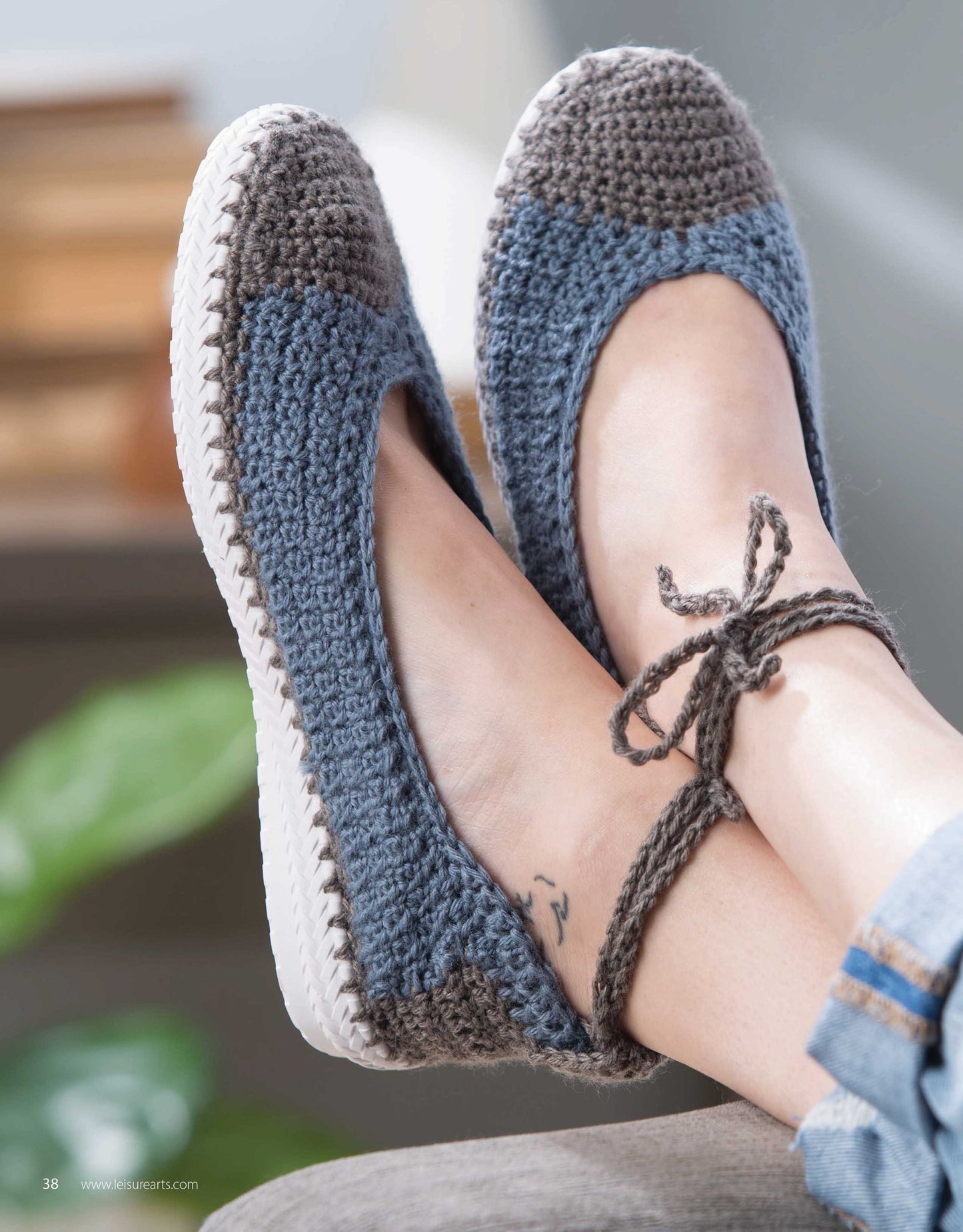 A close-up shows a person wearing Leisure Arts Crochet Shoes in blue and gray with white soles. The stylish slippers feature ankle ties and resemble chic crochet footwear; a small tattoo is visible above the foot.