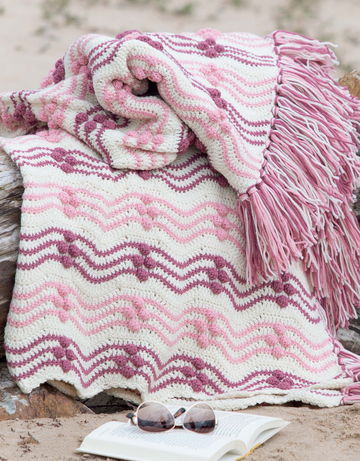 The Restful Afghans Crochet blanket by Leisure Arts, featuring a pink, purple, and white chevron design with fringe, is draped over a log on the sand. An open book and round sunglasses are placed next to it.
