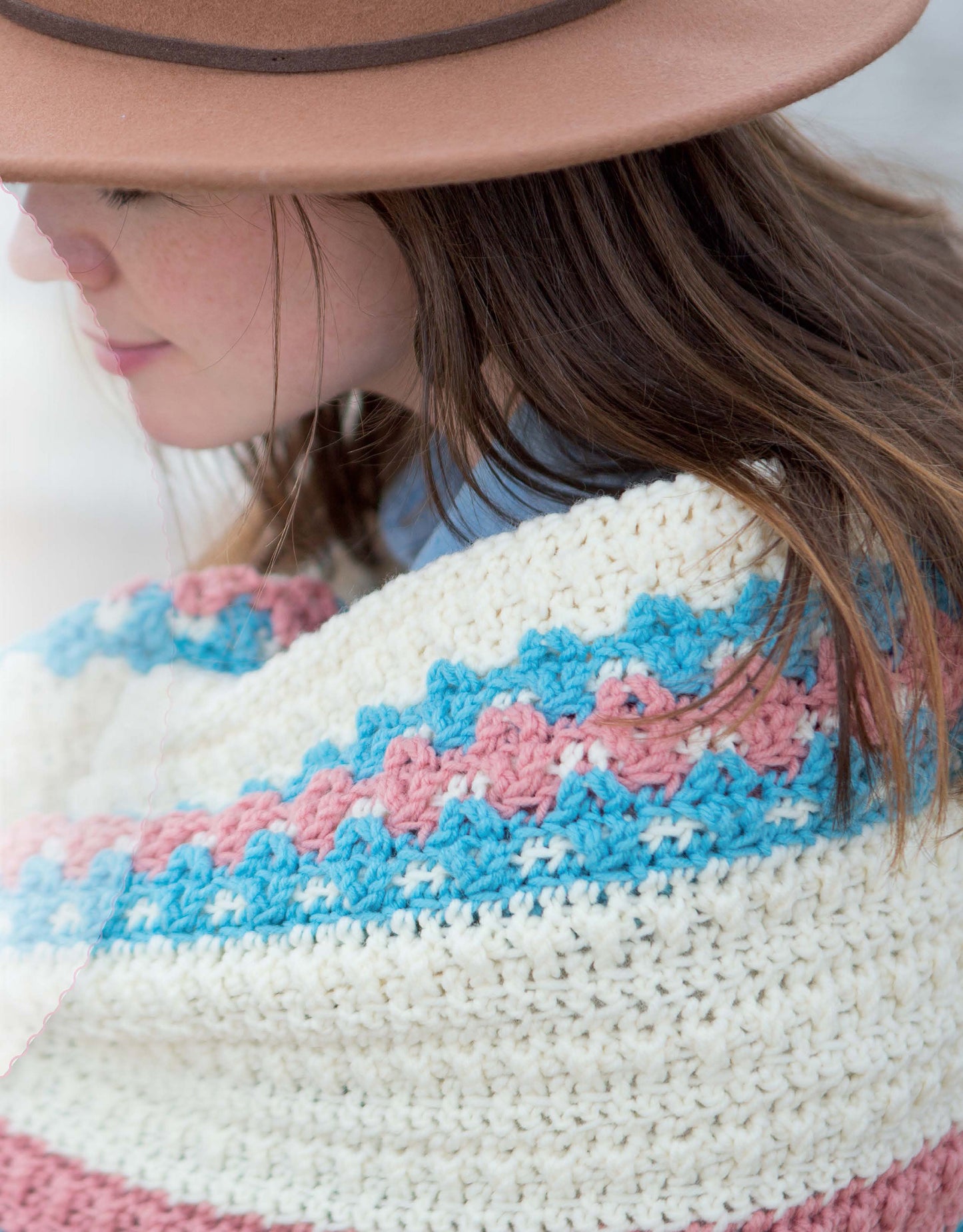 A woman with long brown hair and a tan hat smiles, wrapped in a blanket made from Leisure Arts’ Restful Afghans Crochet. Cream, blue, and pink stripes showcase beginner-friendly patterns using medium weight yarn.
