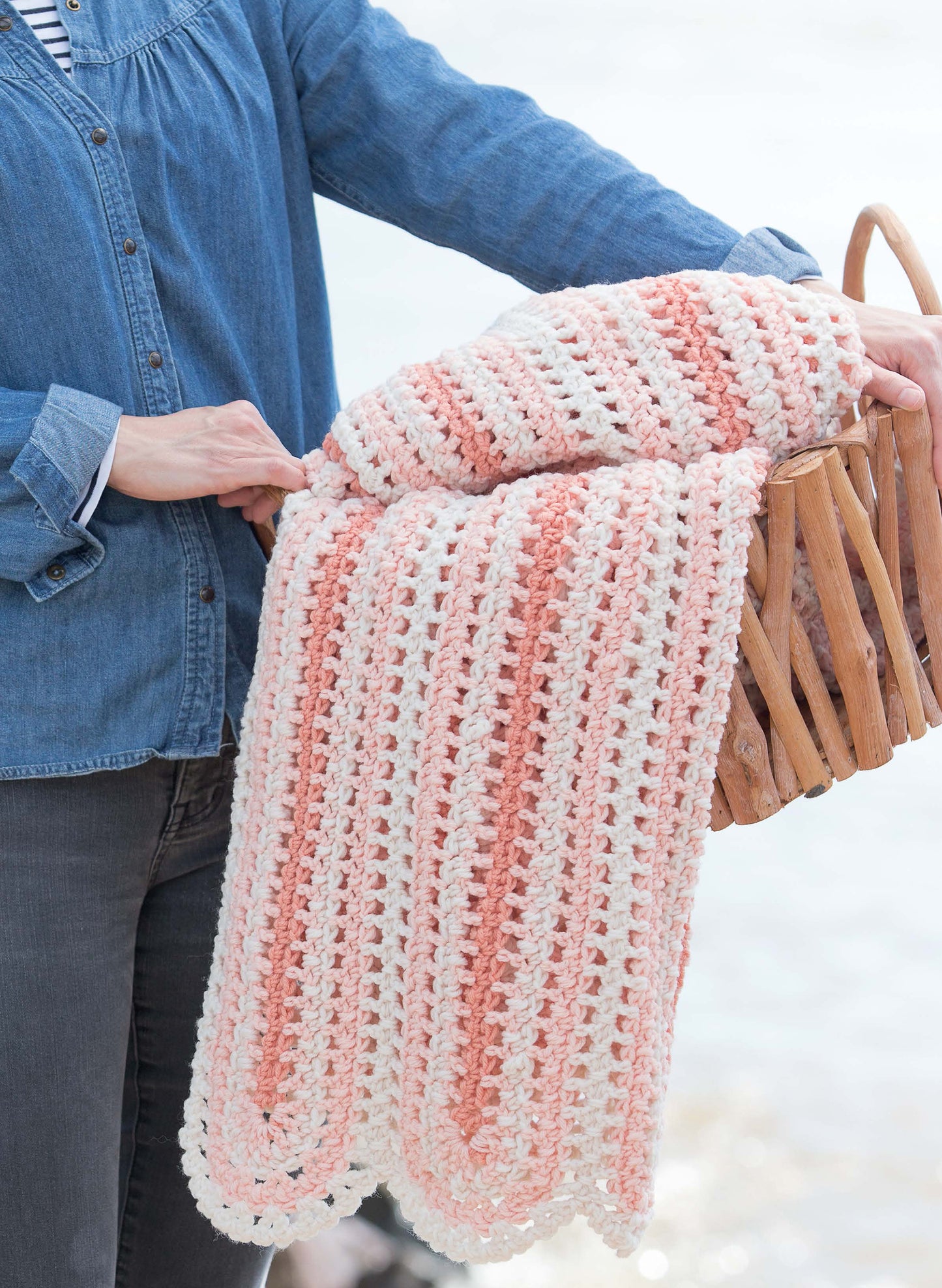 A person in a denim shirt holds a wooden basket with a pink and white striped blanket from the Leisure Arts Restful Afghans Crochet collection, featuring an airy stitch pattern—ideal for beginner crochet projects with medium weight yarn.
