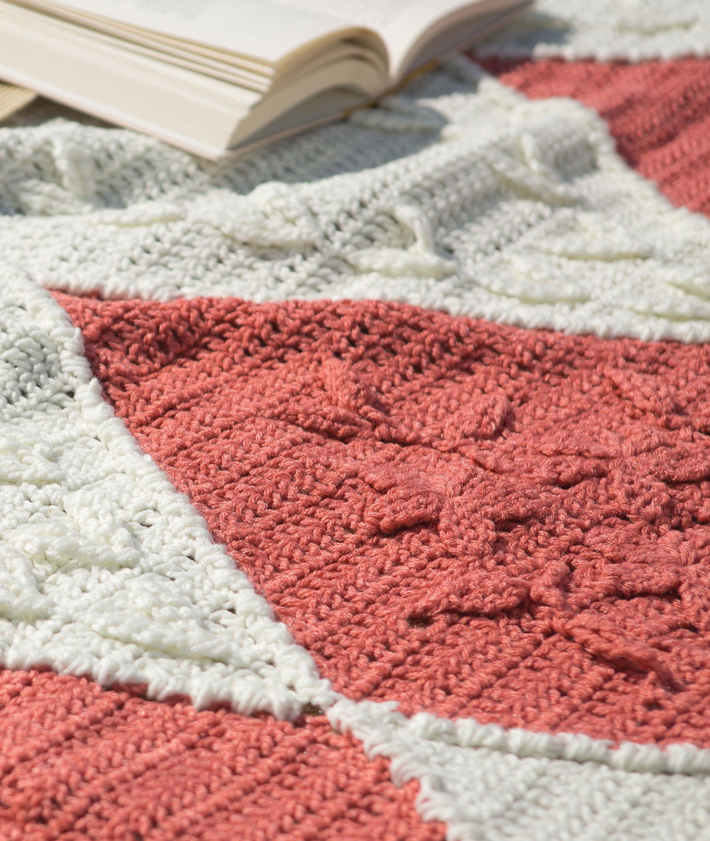 A close-up of a Restful Afghans Crochet blanket by Leisure Arts, made with medium weight yarn in textured white and pink squares, is shown lying on a surface with an open book in the background.