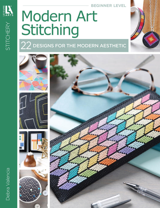 Modern Art Stitching Digital Download by Leisure Arts features 22 geometric cross-stitch projects for all skill levels on 14 count Aida, shown with eyeglasses, pen, notebook, plant, and colorful stitched items.