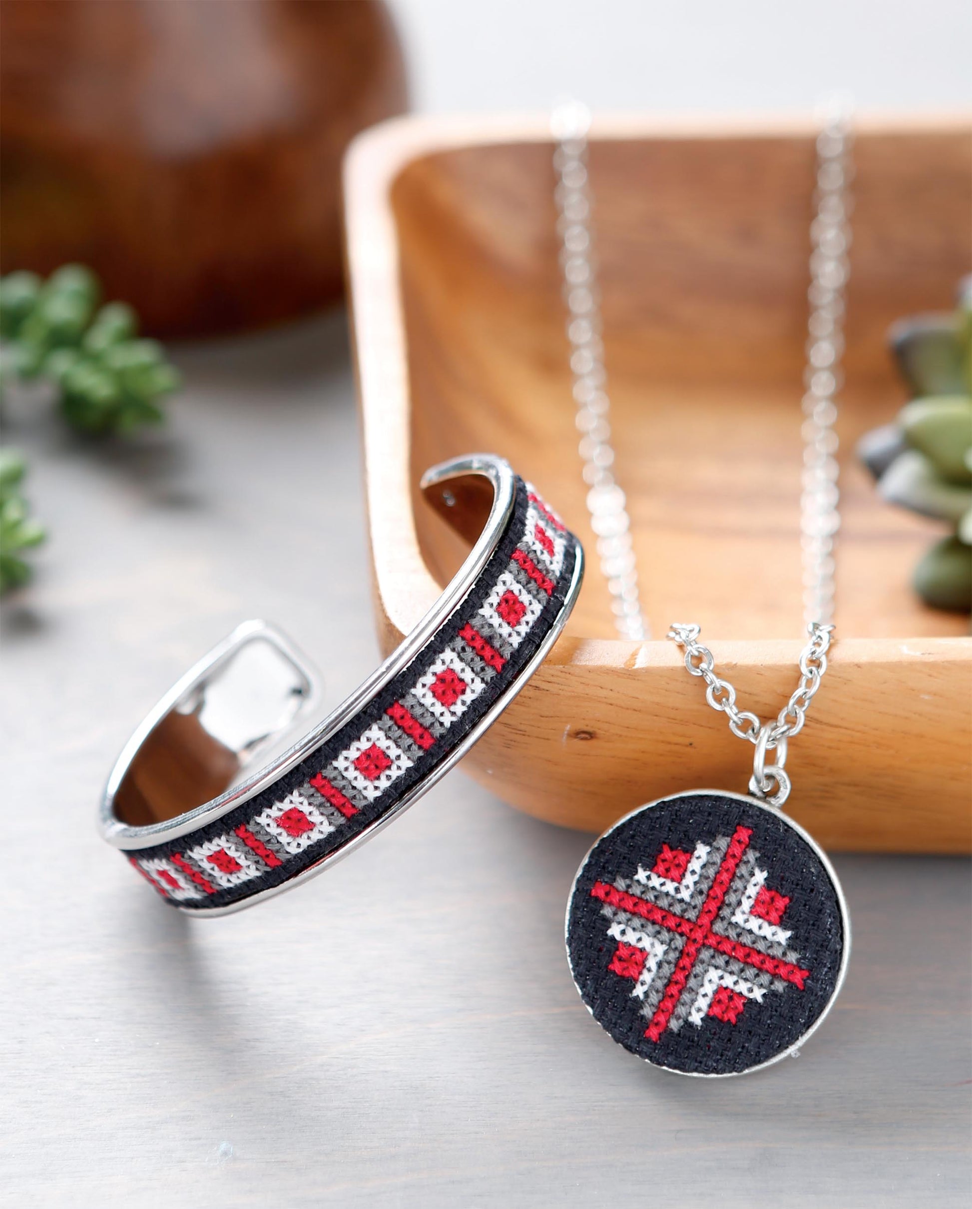 A silver bracelet and a round pendant necklace display red, black, and white cross stitch designs from the Leisure Arts Modern Art Stitching Digital Download, shown on a light surface near a wooden tray and succulents.