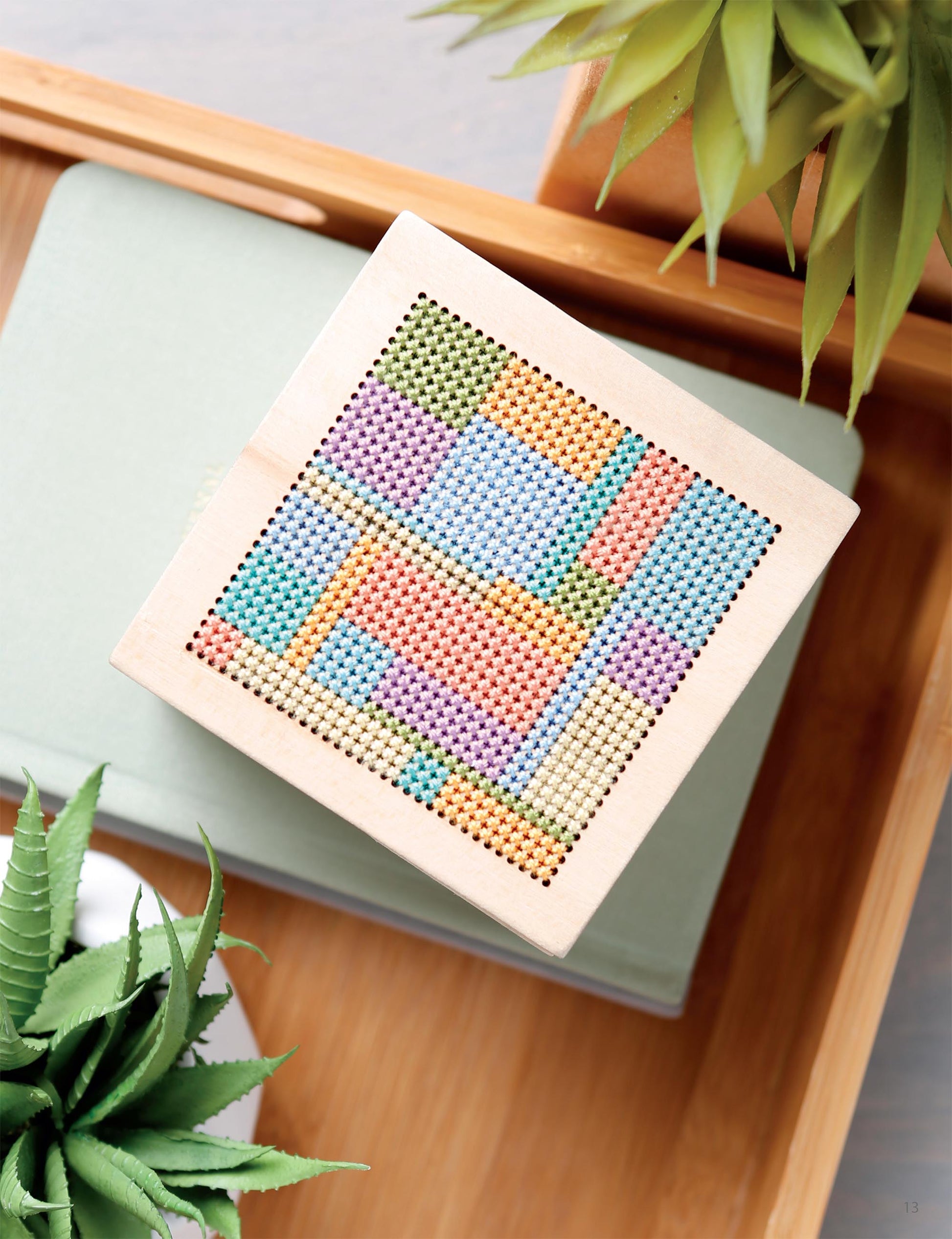 The Modern Art Stitching Digital Download by Leisure Arts features a colorful geometric cross-stitch pattern on 14 count Aida—ideal inspiration for beginner to advanced cross stitchers, displayed with succulents and a closed book.
