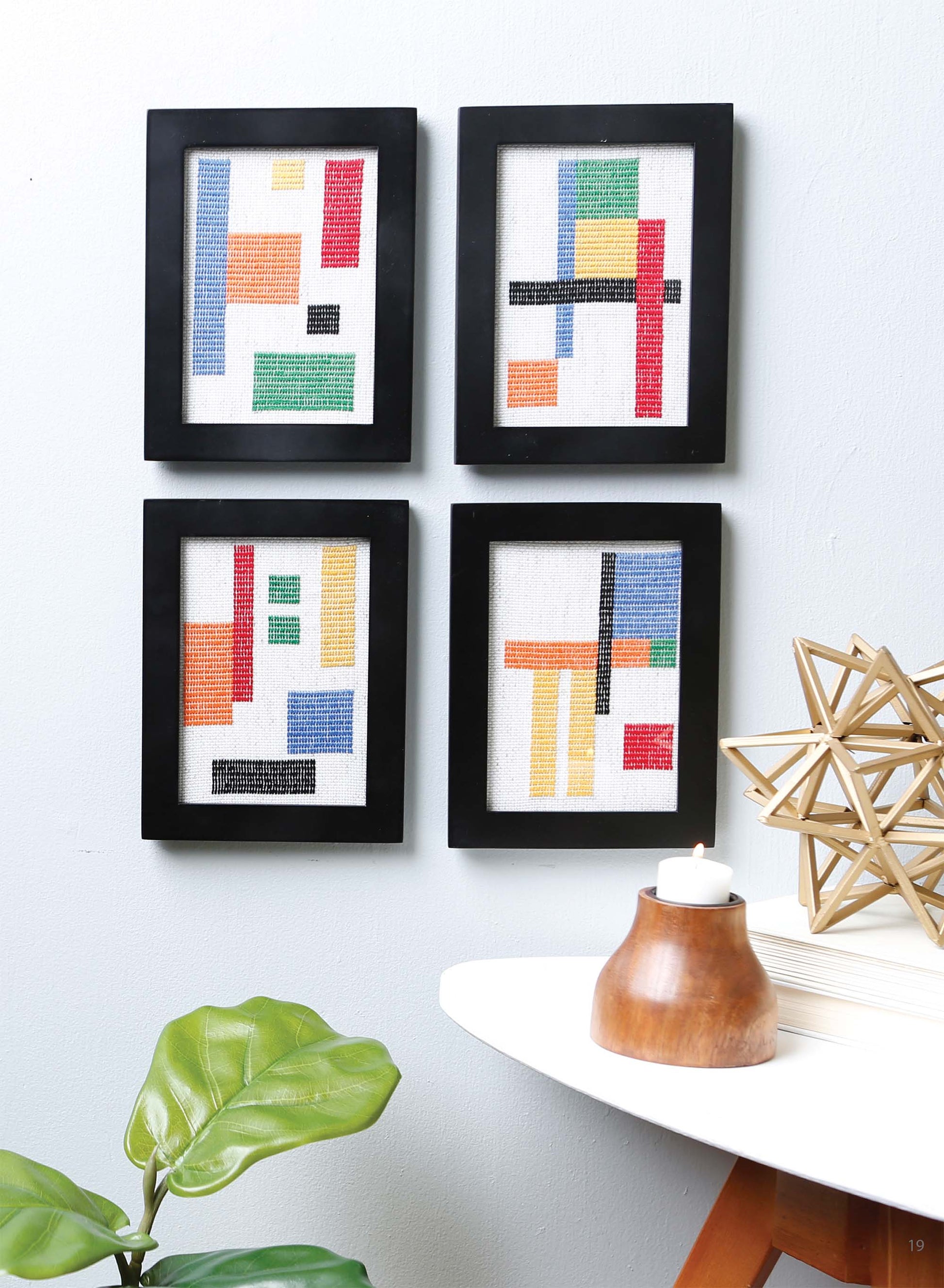 Four black-framed Modern Art Stitching Digital Download artworks by Leisure Arts, featuring colorful geometric patterns, hang above a white table displaying cross stitch projects, a candle, books, a gold sculpture, and a leafy plant.