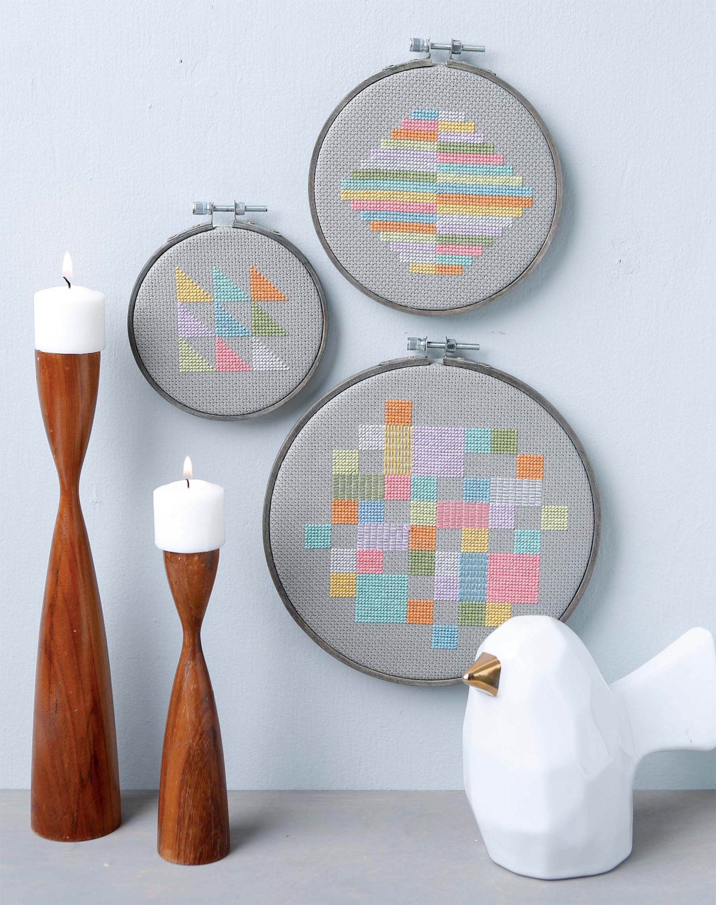 Three Modern Art Stitching Digital Downloads by Leisure Arts, featuring colorful geometric embroidery patterns for all skill levels, are displayed on a light gray wall with candles and a white ceramic bird with a gold beak.