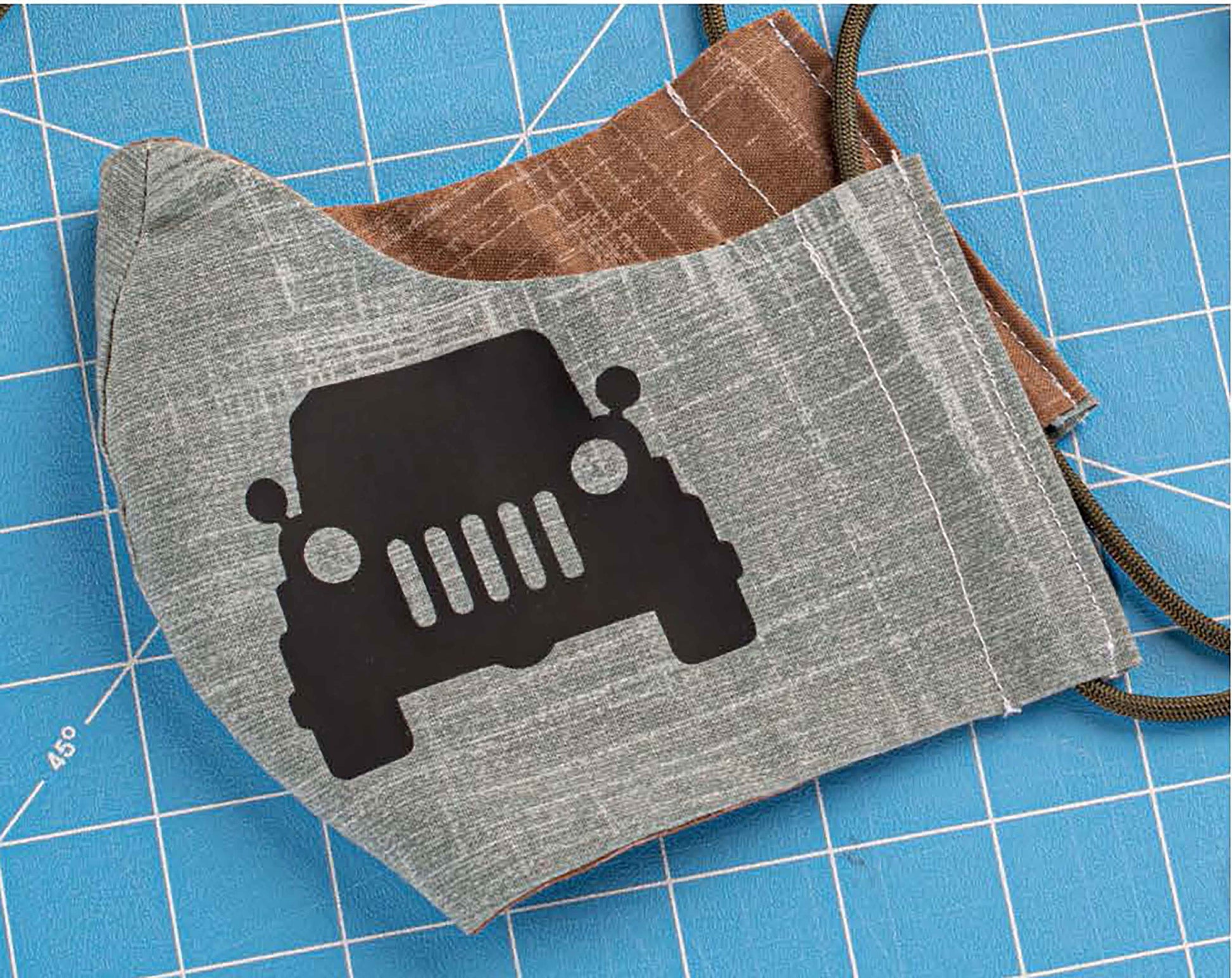 A cloth face mask featuring a black jeep silhouette, inspired by "Breathe Easy 2—30 Embellishments for Adding Style to Any Mask" from Leisure Arts, rests on a blue cutting mat with white grid lines. The mask has brown straps and a brown lining.