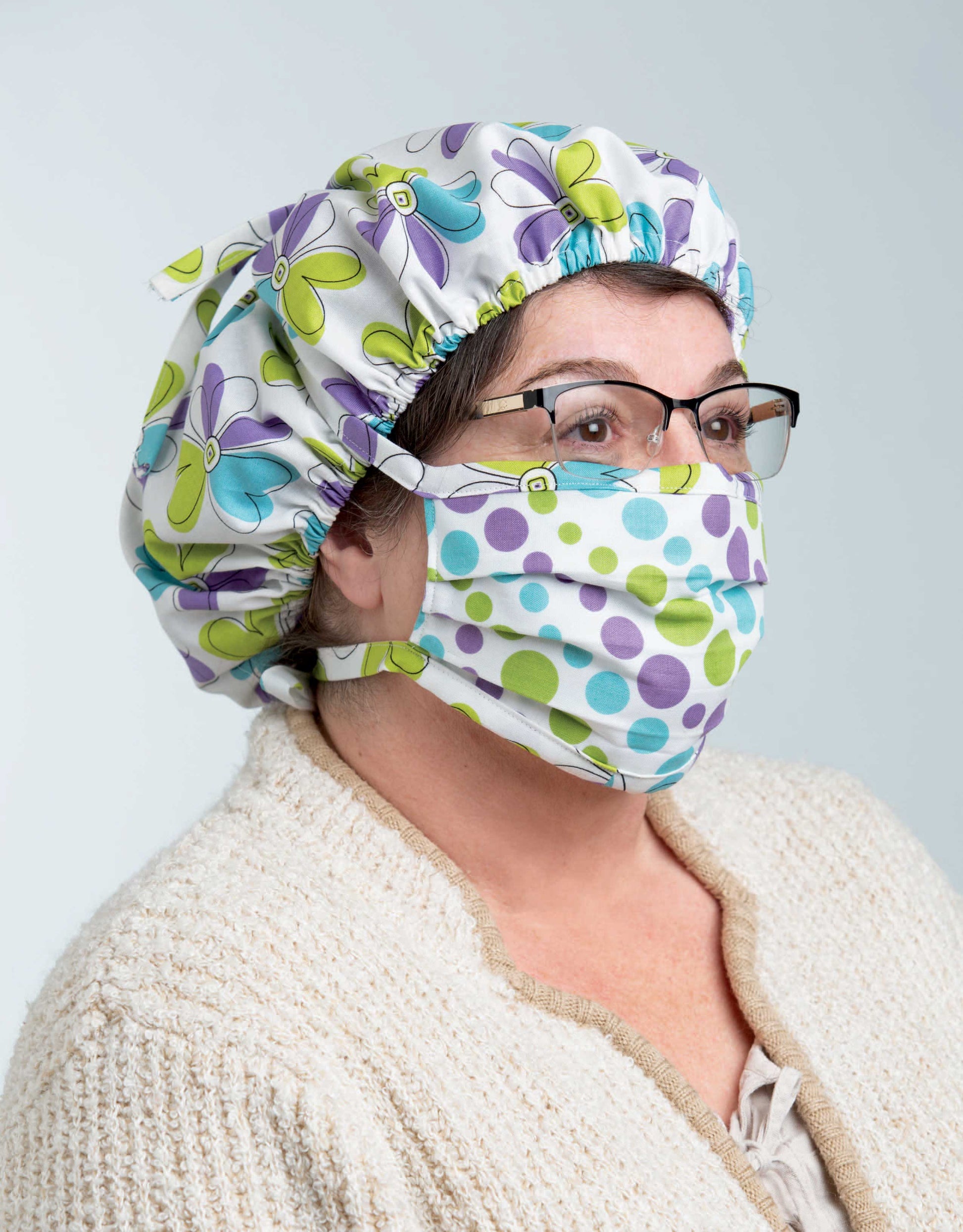 A person wears glasses, a beige textured sweater, a floral scrub cap, and a polka-dot fabric mask made using "Breathe Easy—8 Simple Sew and Applique Particle Mask and Cap Projects" by Leisure Arts, standing against a light background.