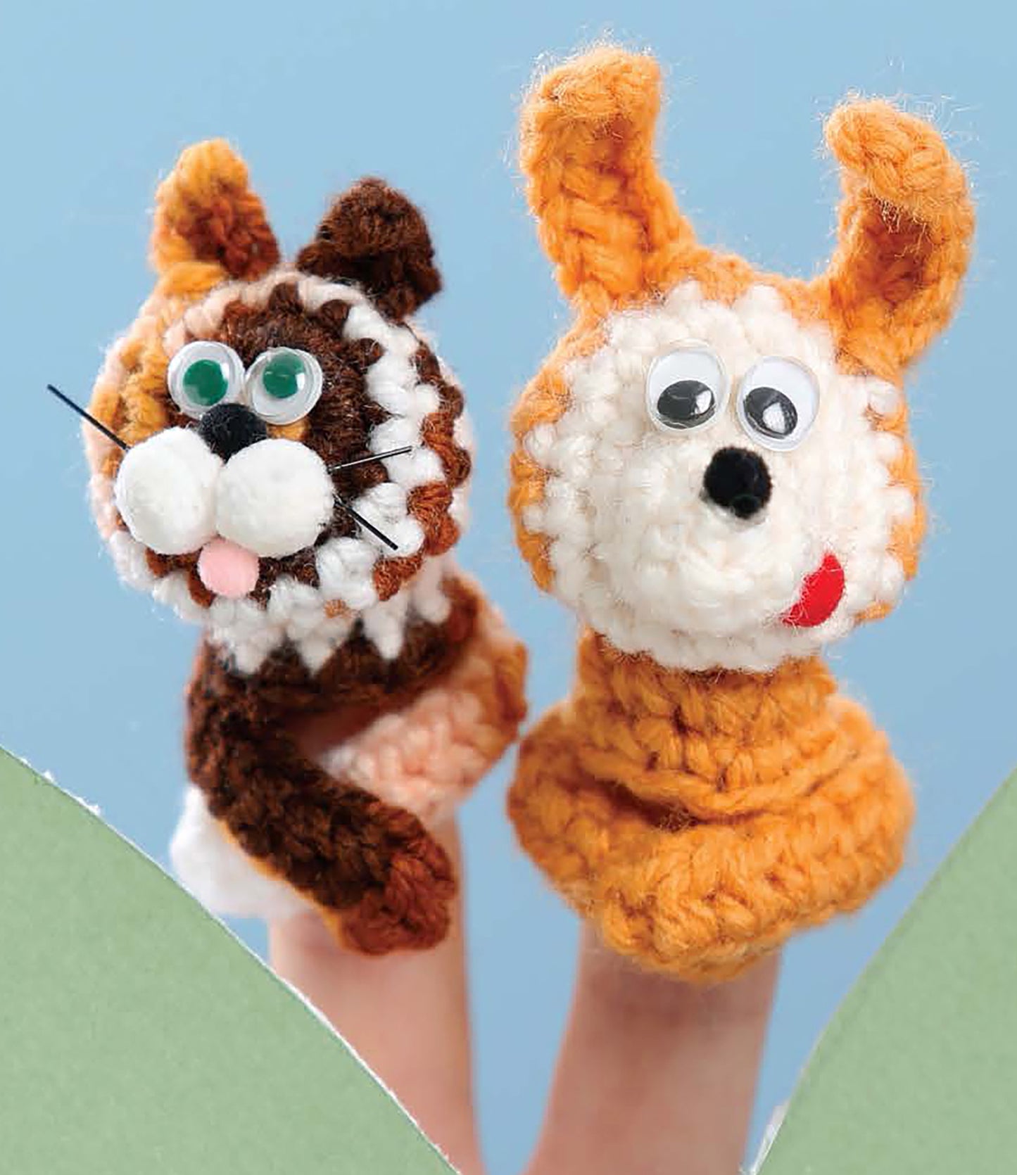 Two fingers display "Finger Puppets Our Town" by Leisure Arts: a brown-and-white cat and an orange-and-white dog, each with googly eyes and pink tongues, set against a blue background. Ideal for beginner crochet enthusiasts.