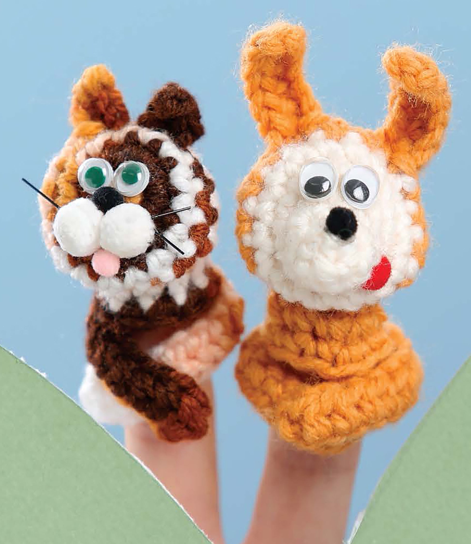 Two fingers display "Finger Puppets Our Town" by Leisure Arts: a brown-and-white cat and an orange-and-white dog, each with googly eyes and pink tongues, set against a blue background. Ideal for beginner crochet enthusiasts.