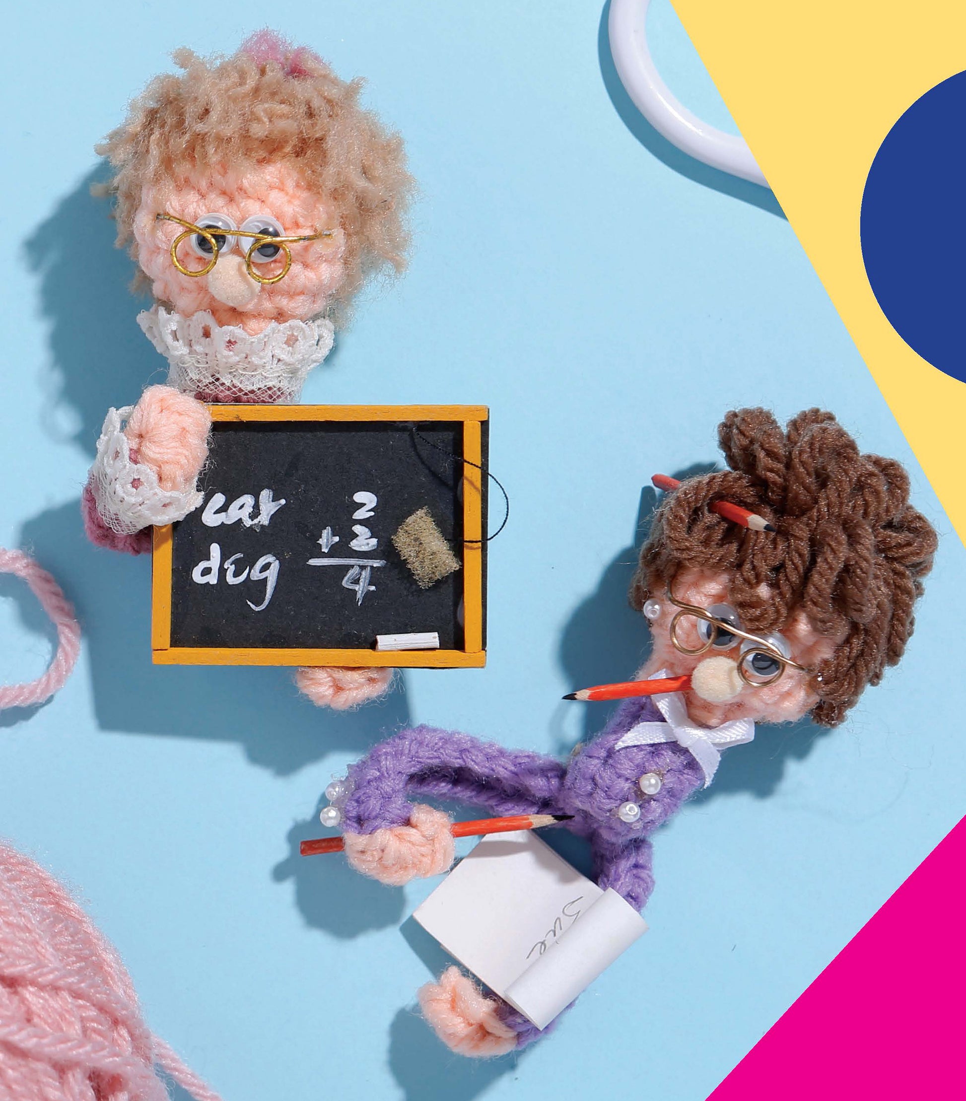Two "Finger Puppets Our Town" by Leisure Arts wear glasses and hold school items—one with a math chalkboard, the other with a notepad and pencil—set on a pastel blue background featuring yellow and pink geometric shapes.