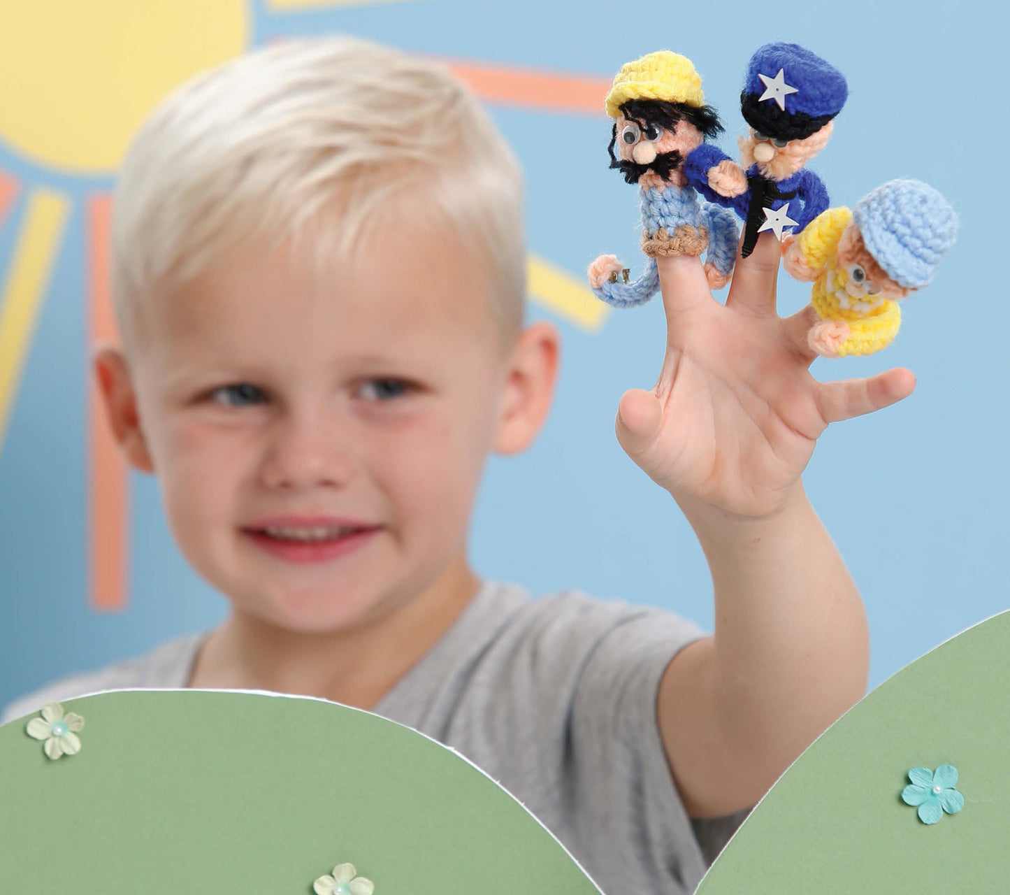 A young blonde boy displays three "Finger Puppets Our Town" by Leisure Arts—blue and yellow characters—on his fingers, with a colorful sun-and-flowers background.