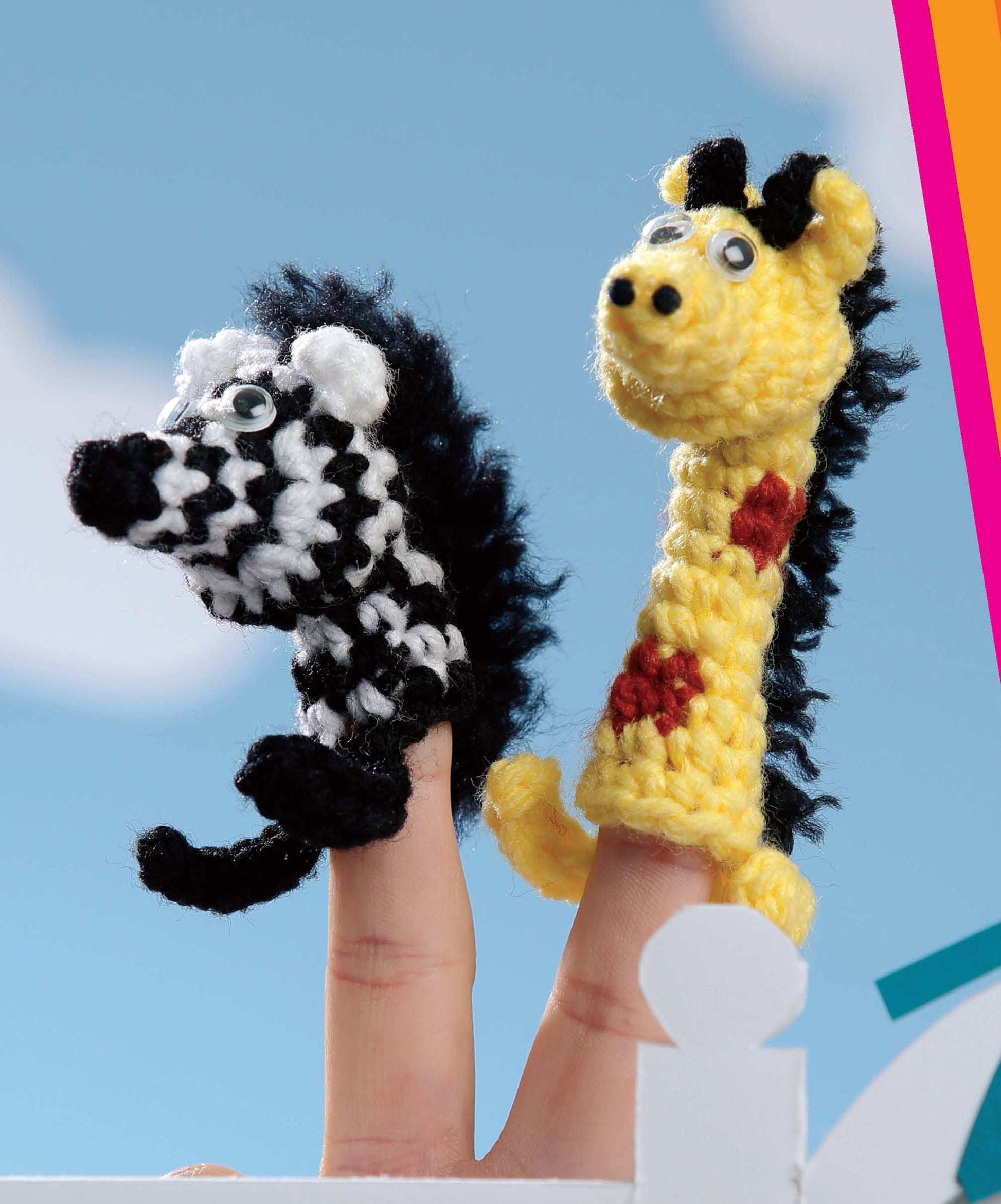 Two fingers wear Leisure Arts' Finger Puppets At The Zoo—a zebra and giraffe, both cute crochet favorites with googly eyes. Perfect beginner projects, they're shown against a blue sky background.