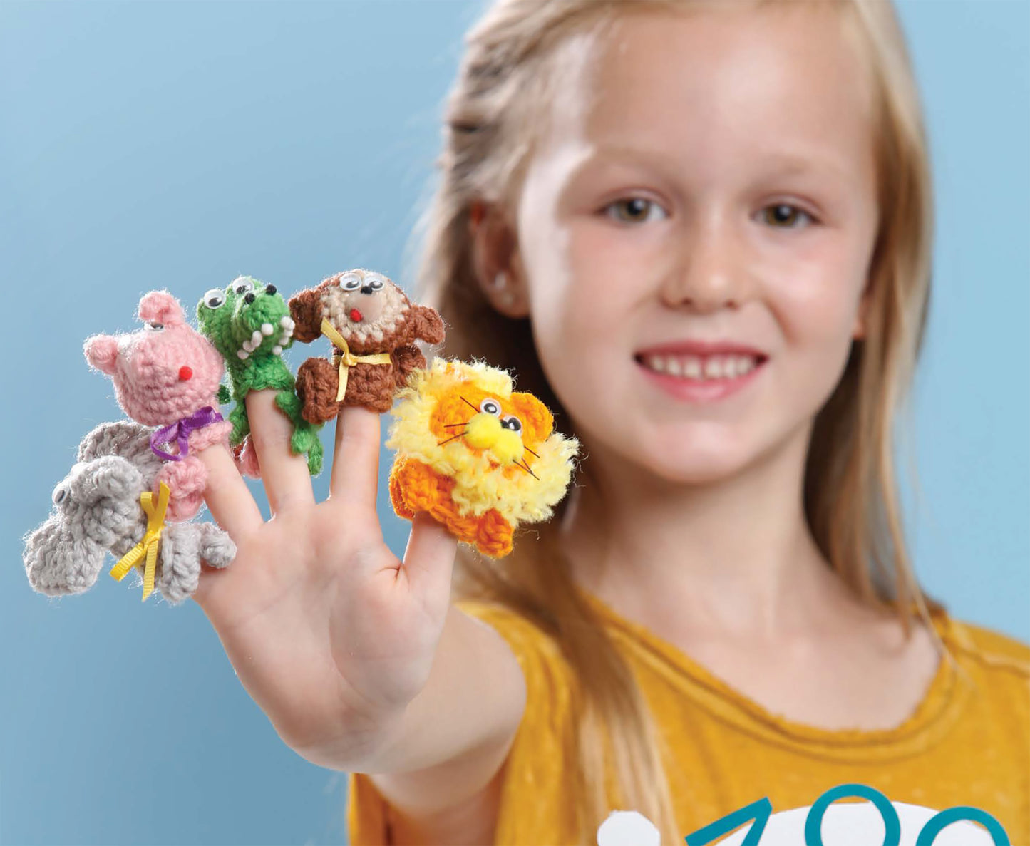 A smiling young girl displays her hand with each finger adorned by a different zoo animal from Leisure Arts’ "Finger Puppets At The Zoo" crochet pattern—the perfect inspiration for beginner crochet projects against a blue background.