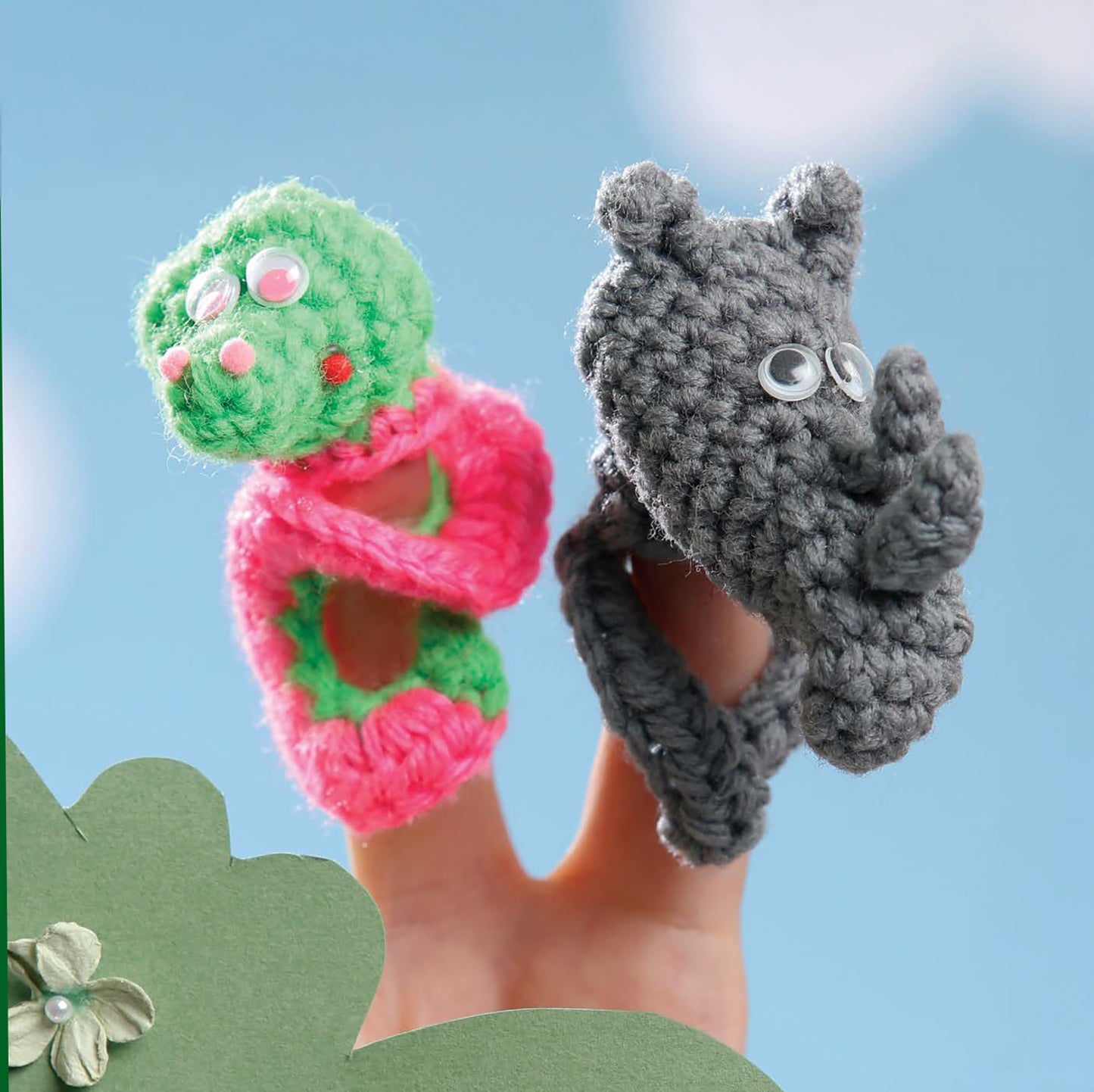 Two fingers display Leisure Arts' "Finger Puppets At The Zoo": a green and pink smiling creature and a gray bear with googly eyes, set on a blue background with clouds—great inspiration for beginner crochet or zoo animal projects.