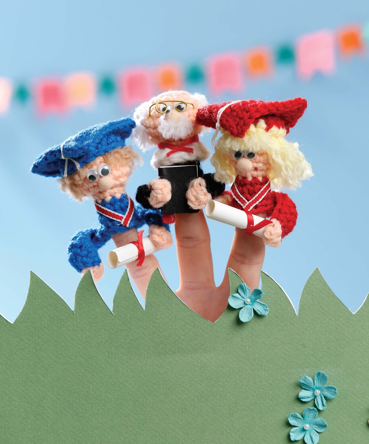 Finger Puppets Celebration by Leisure Arts features three beginner-level crocheted finger puppets resembling graduates in blue and red caps with diplomas, plus one with glasses and a book, displayed above green grass with a colorful background.