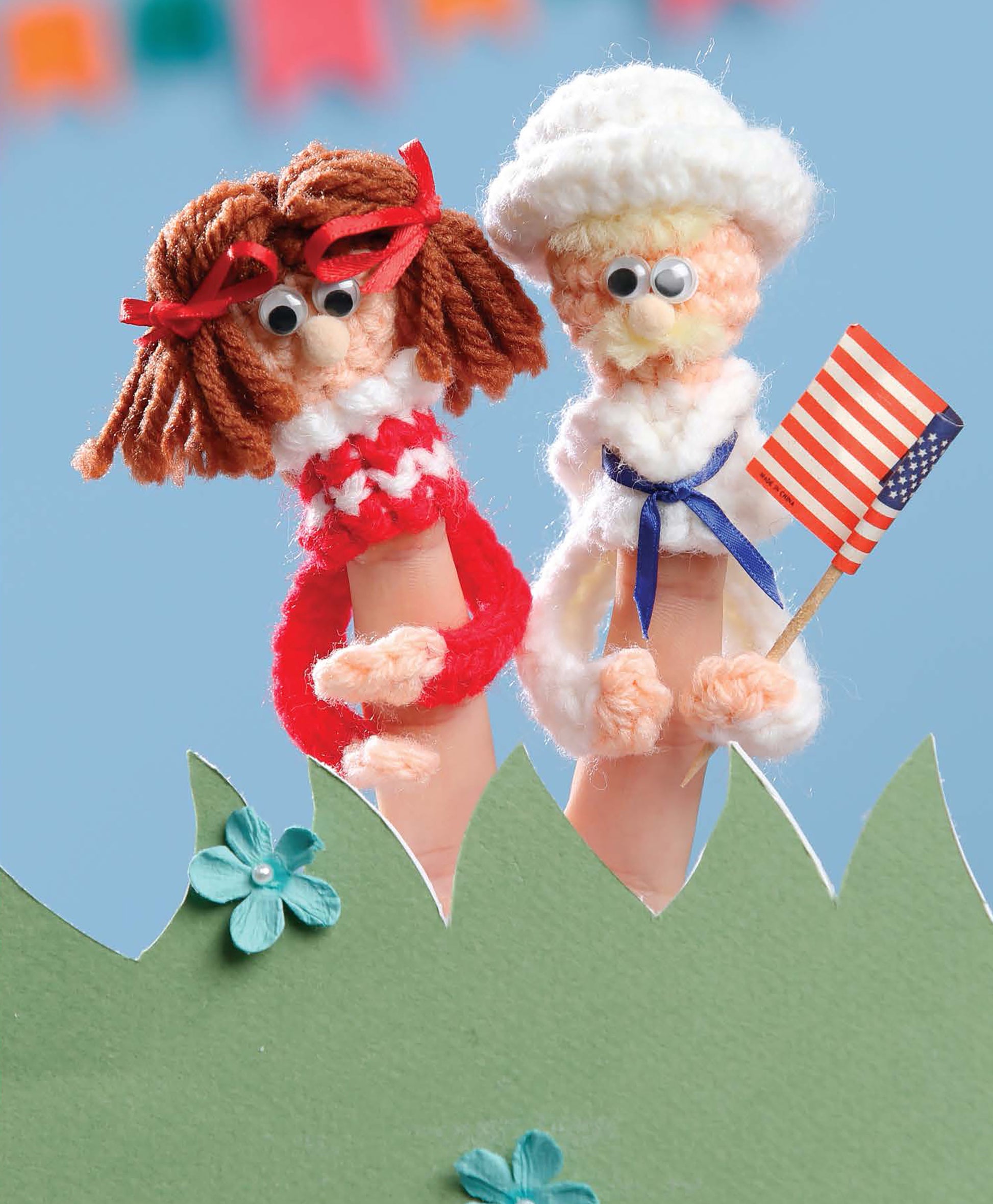 Finger Puppets Celebration by Leisure Arts features two beginner crochet yarn puppets with googly eyes and pipe cleaner arms—one with red hair, the other in a white outfit holding an American flag—standing above green paper grass and blue flowers.
