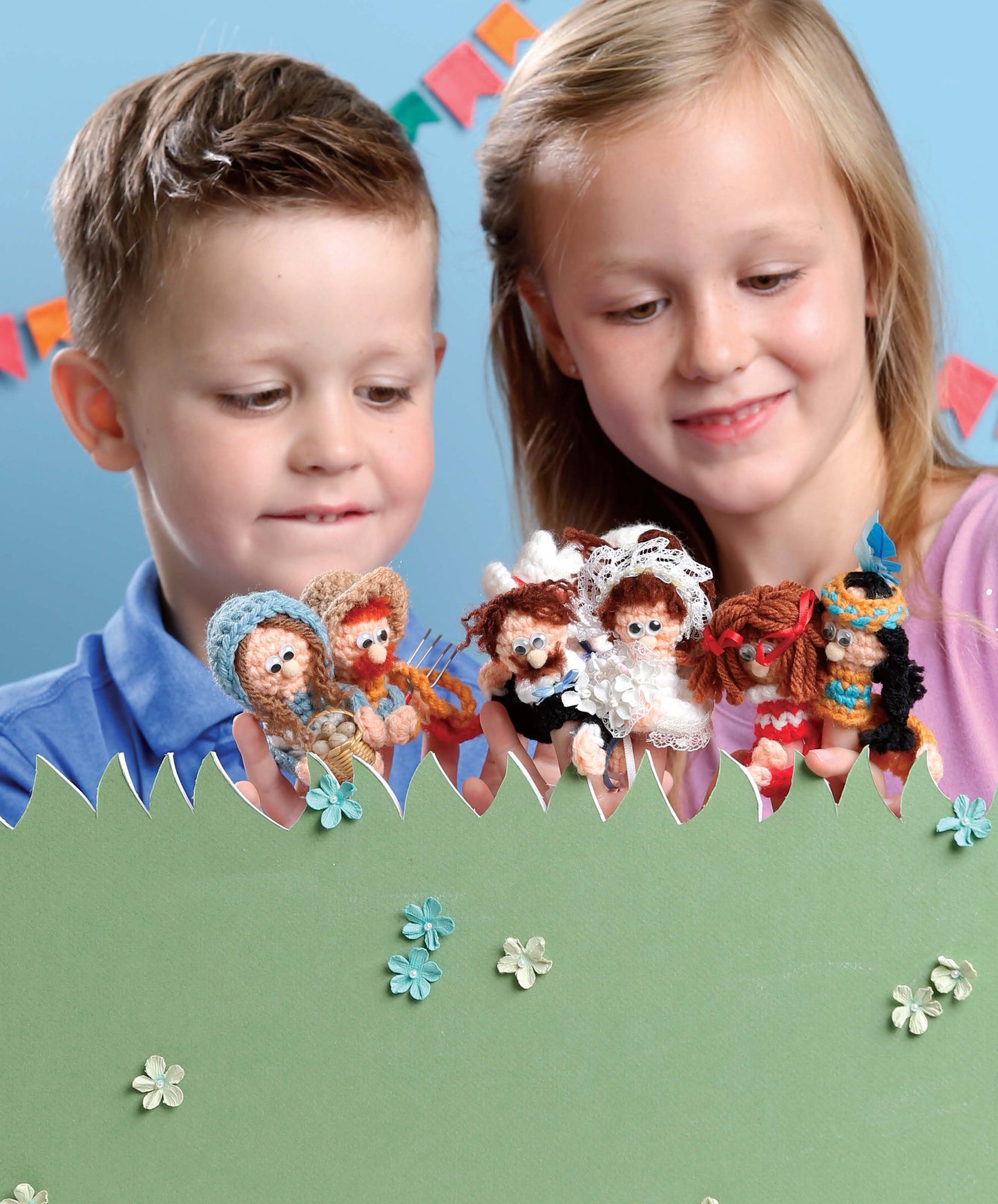 Two children smile and hold colorful Finger Puppets Celebration by Leisure Arts over a green paper hedge with blue flowers, set against a blue background and festive bunting—perfect for sparking creativity and beginner crochet fun.