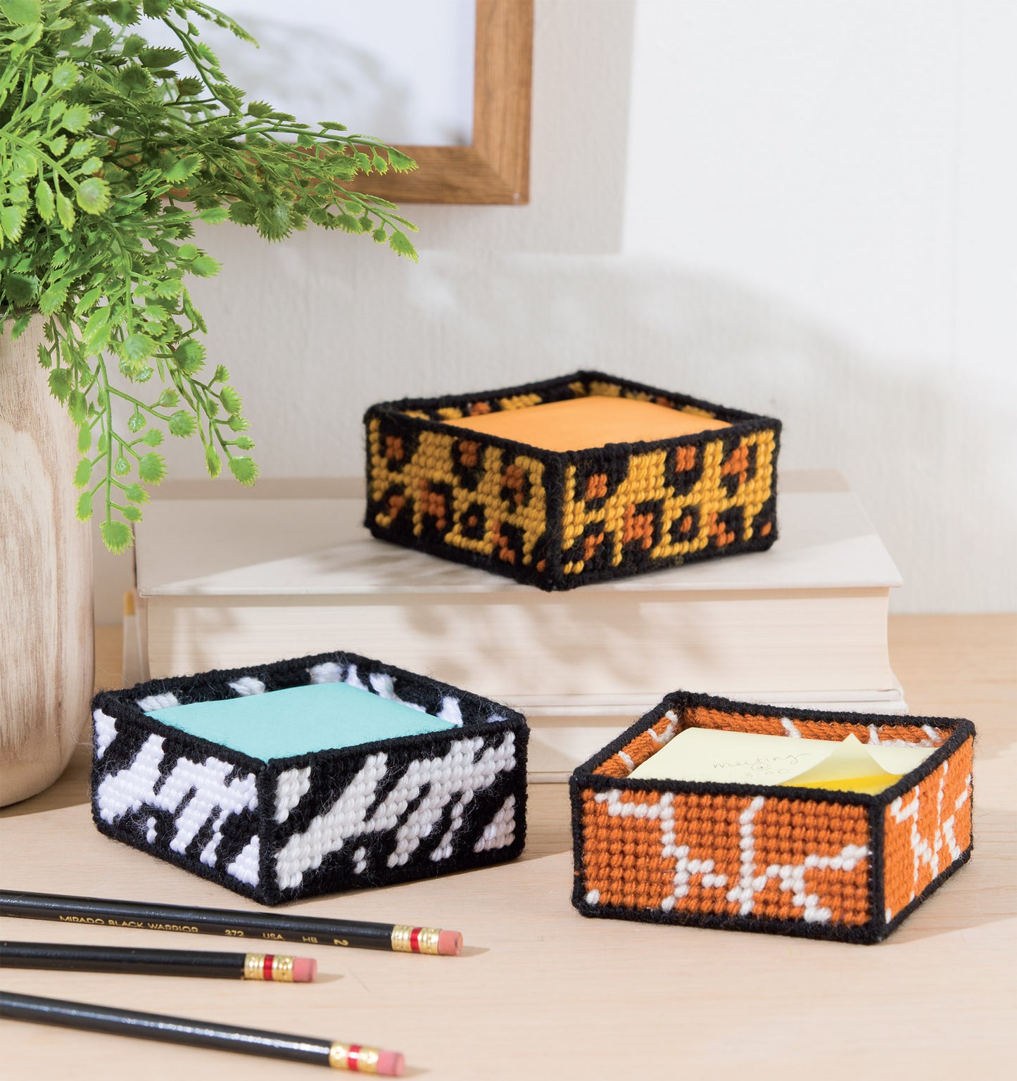 Leisure Arts’ Wild for Style—Plastic Canvas Desk Sets features three square, open-top, handwoven animal-print boxes holding sticky notes on a desk with pencils, a book, and plant—ideal for creative home accessories.