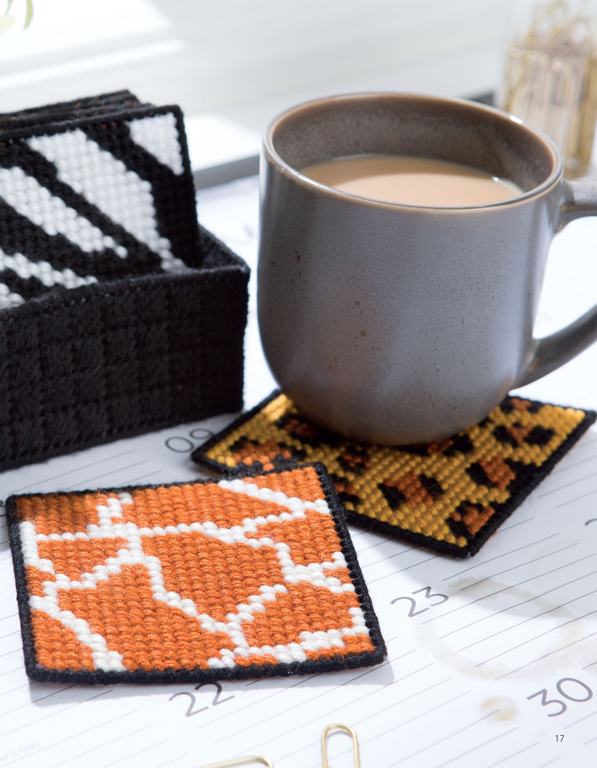 A gray mug of coffee sits on a stitched coaster atop an open planner, with more geometric-patterned plastic canvas desk projects from Wild for Style—Plastic Canvas Desk Sets by Leisure Arts visible nearby, plus a black box with white and black design.