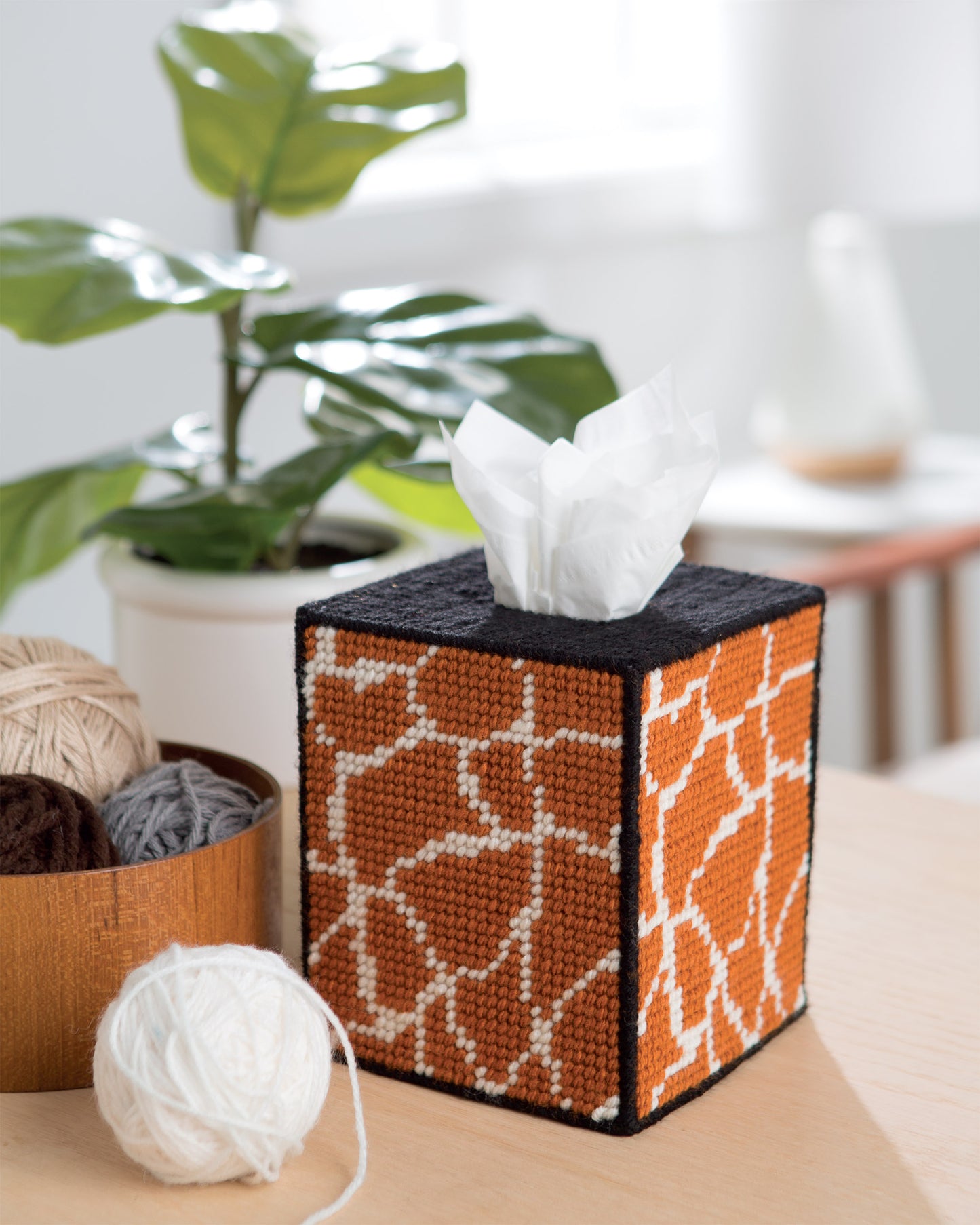 On a table sits a tissue box from Leisure Arts’ "Wild for Style—Plastic Canvas Desk Sets and Accessories for Your Home," featuring a hand-stitched giraffe-patterned cover, alongside a wooden bowl of yarn, with greenery and blurred decor in the background.