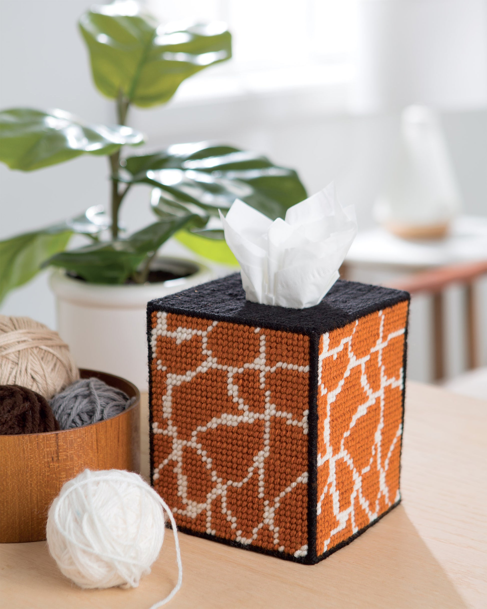 On a table sits a tissue box from Leisure Arts’ "Wild for Style—Plastic Canvas Desk Sets and Accessories for Your Home," featuring a hand-stitched giraffe-patterned cover, alongside a wooden bowl of yarn, with greenery and blurred decor in the background.
