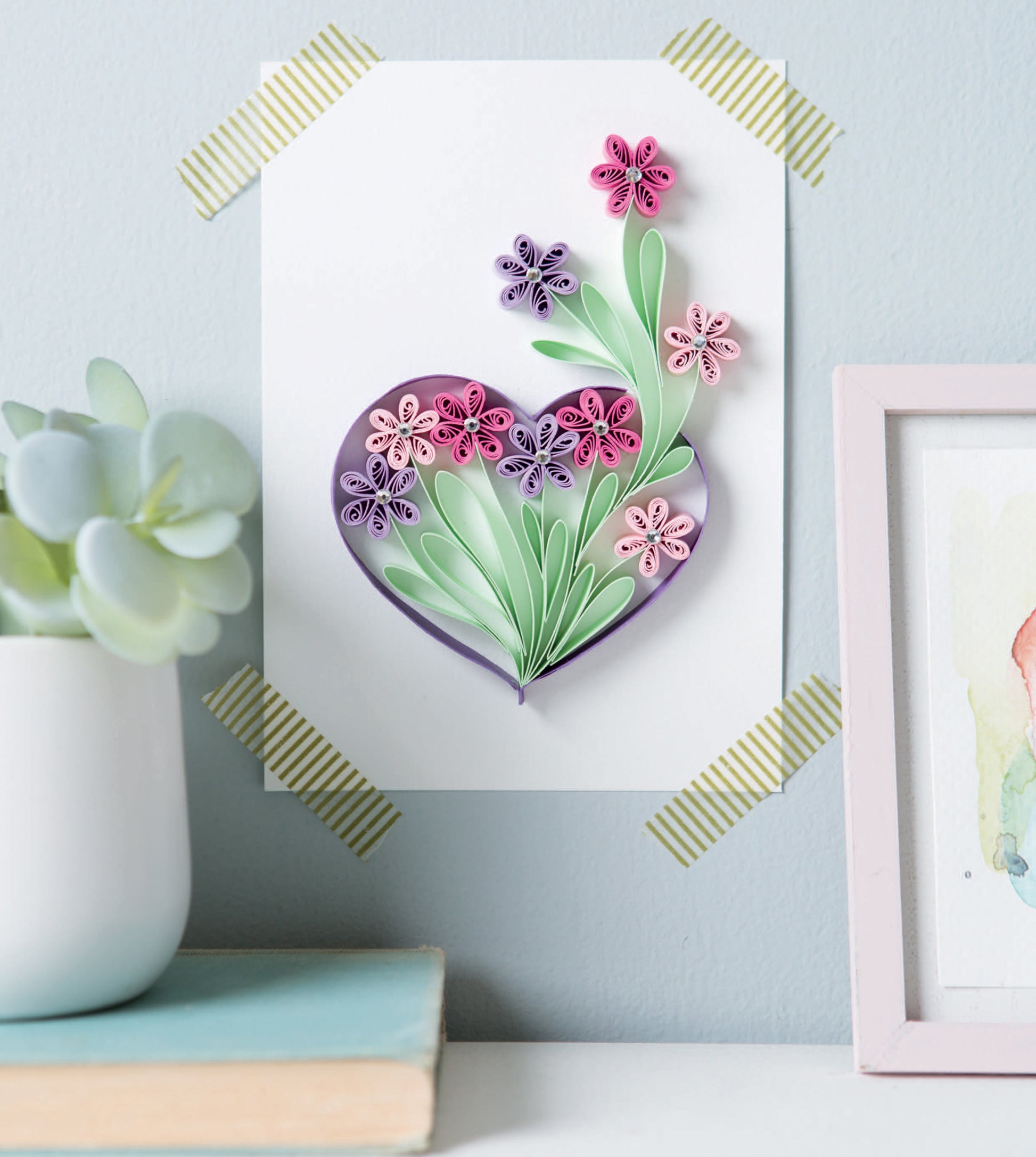 The Quilling for Beginners Digital Download by Leisure Arts shows how to create a heart-shaped paper quilling artwork with pink and purple flowers—perfect home decor. Pair it with a potted succulent and framed watercolor for a lovely craft display.