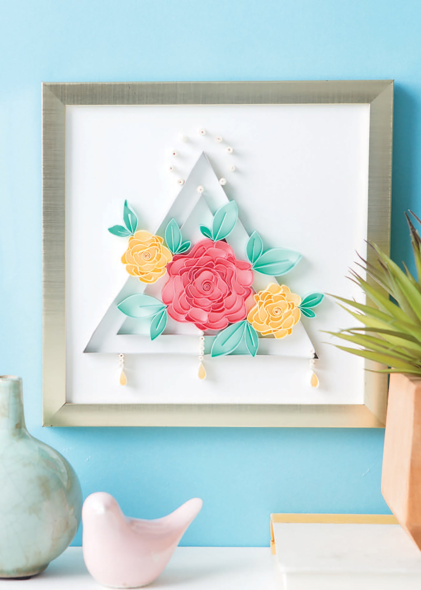Quilling for Beginners Digital Download by Leisure Arts: Create stunning paper quilled triangles with red and yellow flowers and green leaves on a white background—ideal for home decor. Styled with a vase and pink bird in the foreground.