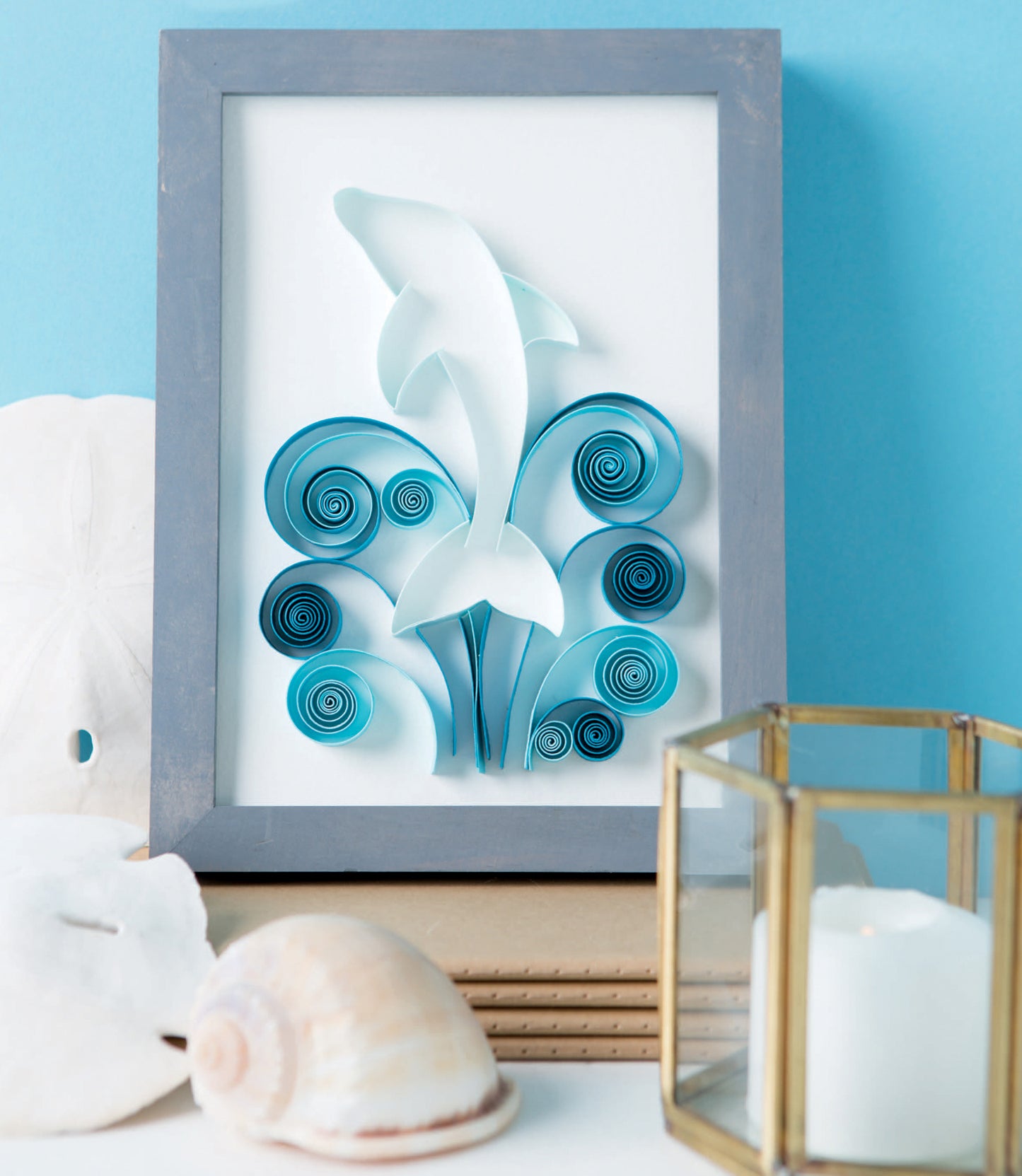 The Quilling for Beginners Digital Download by Leisure Arts features framed dolphin paper art above blue quilled waves, styled with seashells, a sand dollar, and a lit candle—a charming decor piece for craft enthusiasts. Soft blue background included.