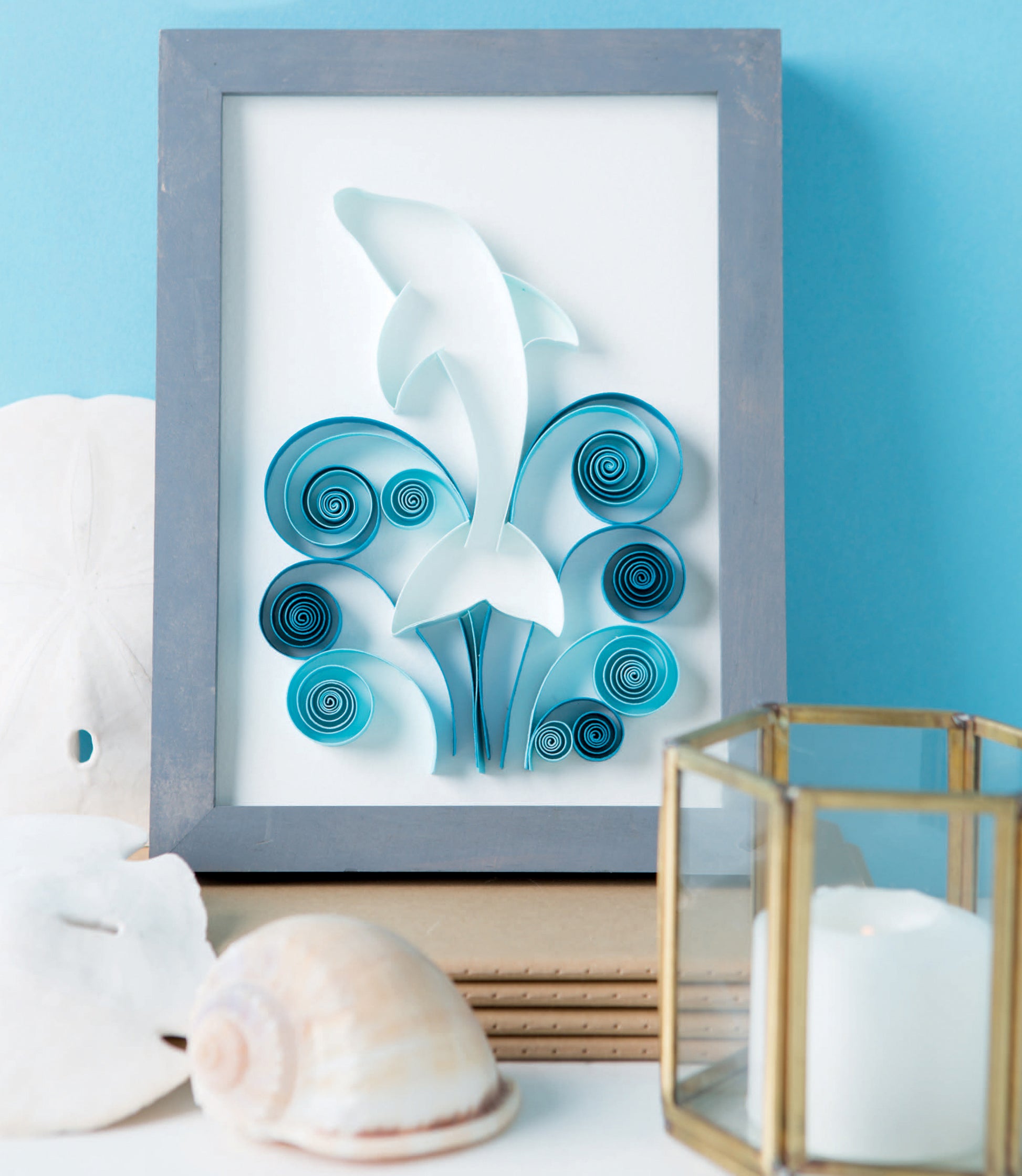 The Quilling for Beginners Digital Download by Leisure Arts features framed dolphin paper art above blue quilled waves, styled with seashells, a sand dollar, and a lit candle—a charming decor piece for craft enthusiasts. Soft blue background included.
