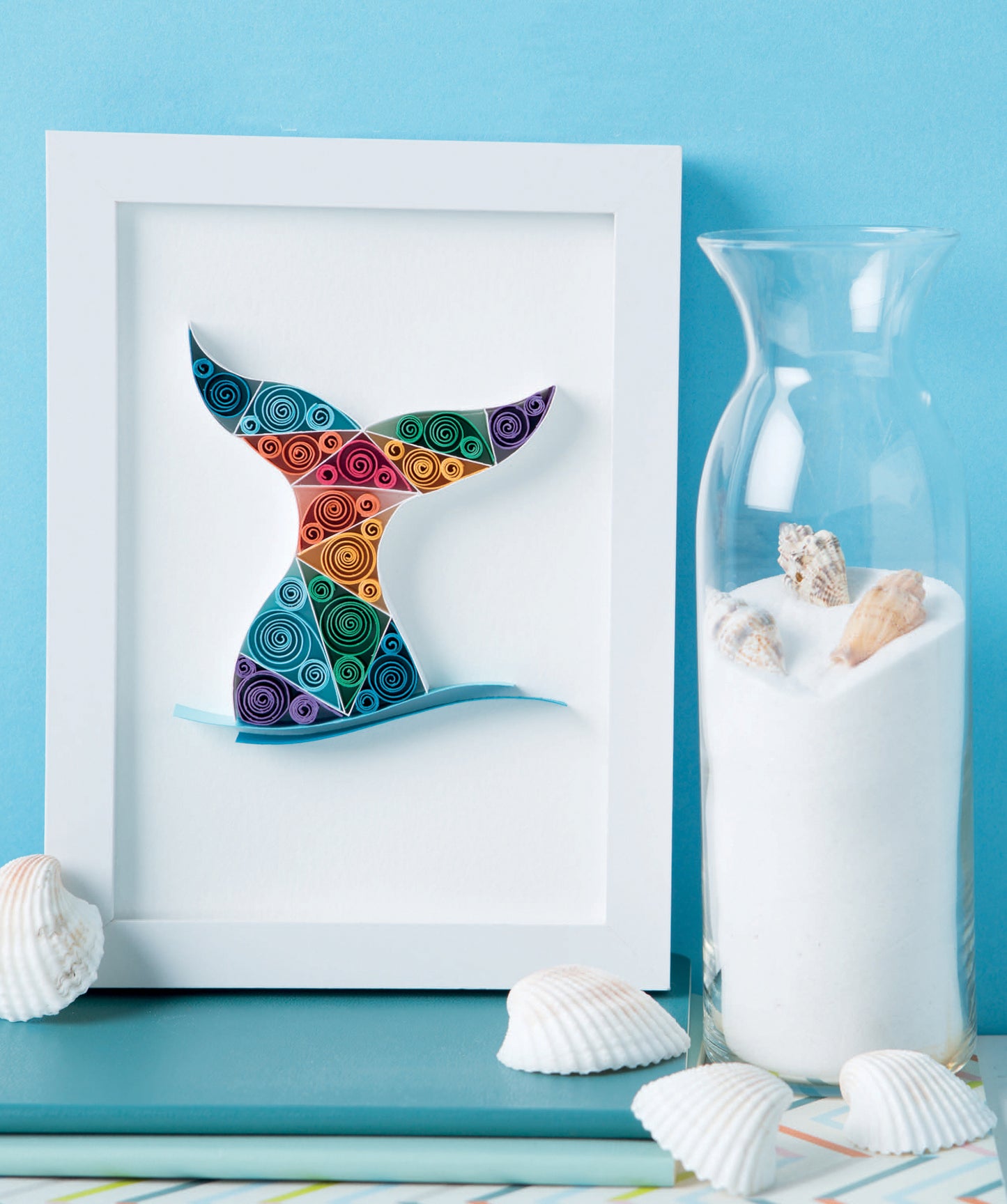 The Quilling for Beginners Digital Download by Leisure Arts features a framed quilled colorful whale tail, displayed next to a vase of white sand and seashells—perfect coastal home decor with scattered seashells on a light blue surface.