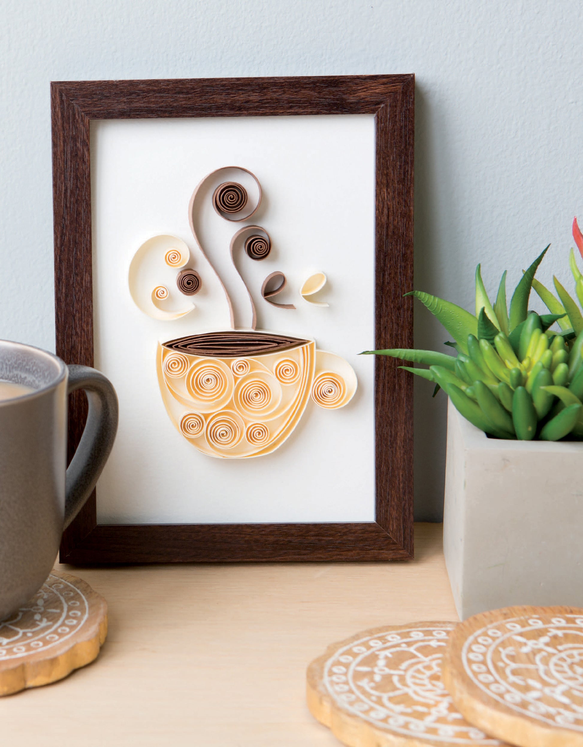 Quilling for Beginners Digital Download by Leisure Arts: framed coffee cup paper quilling art with swirling steam, styled with a mug, coasters, and a succulent—ideal beginner craft decor for your home.