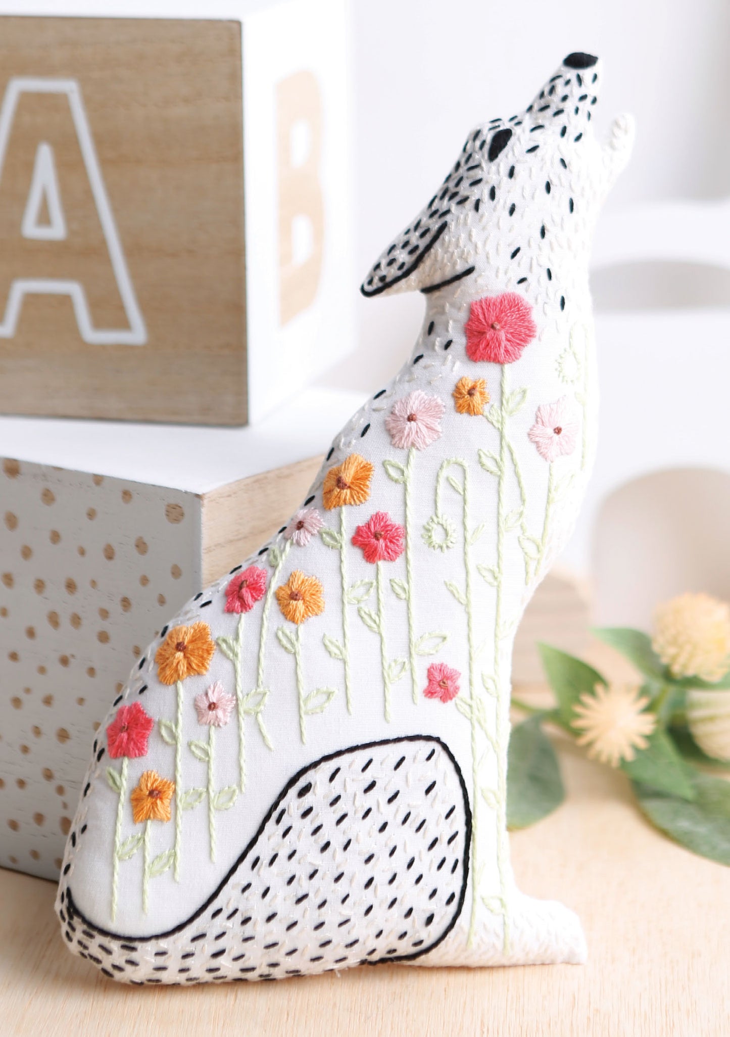 A white coyote plush with embroidered black stitches and colorful flowers, inspired by animal patterns from "Forest Friends—12 Adorable Animals to Embroider by Hand" by Leisure Arts, sits on a light surface with wooden blocks and greenery in the background.