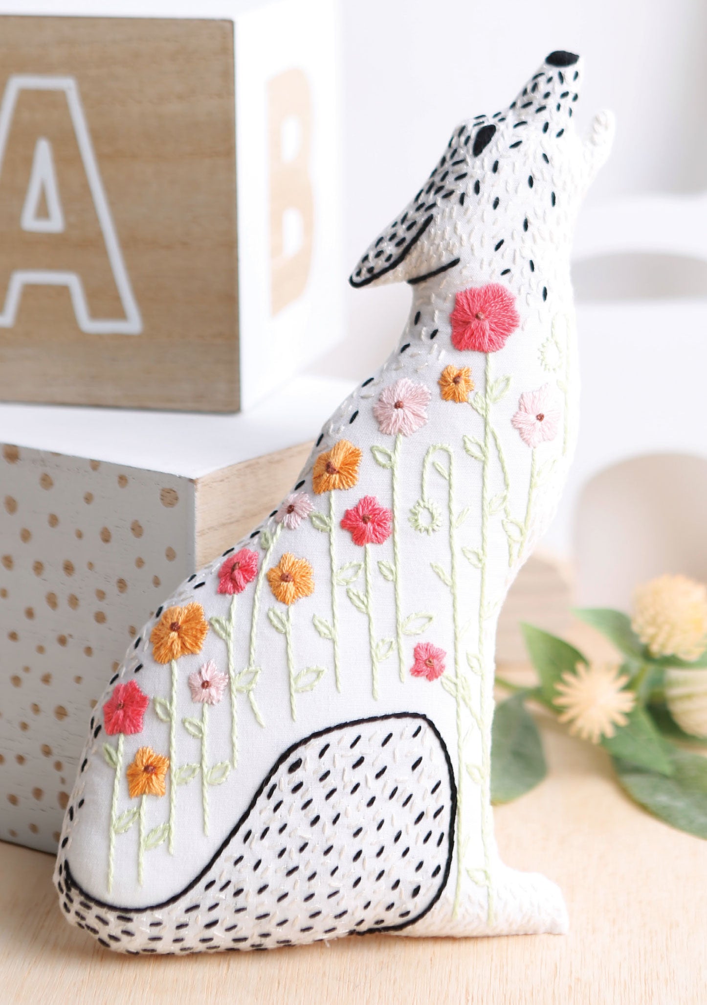 A white coyote plush with embroidered black stitches and colorful flowers, inspired by animal patterns from "Forest Friends—12 Adorable Animals to Embroider by Hand" by Leisure Arts, sits on a light surface with wooden blocks and greenery in the background.