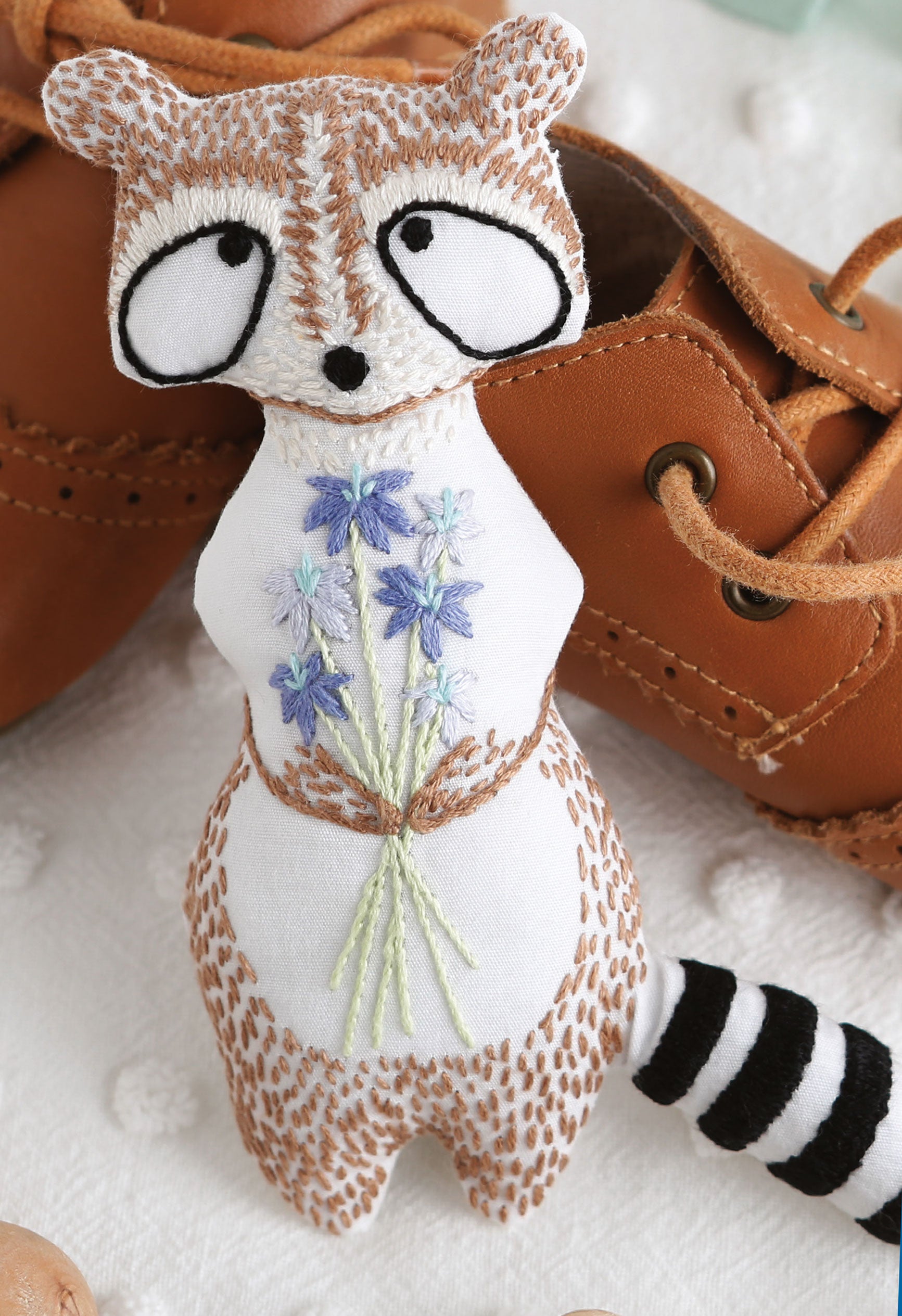 A brown, white, and black hand-embroidered raccoon holding blue flowers—made using the Forest Friends—12 Adorable Animals to Embroider by Hand Digital Download by Leisure Arts—is displayed beside a brown leather shoe on a white textured surface.
