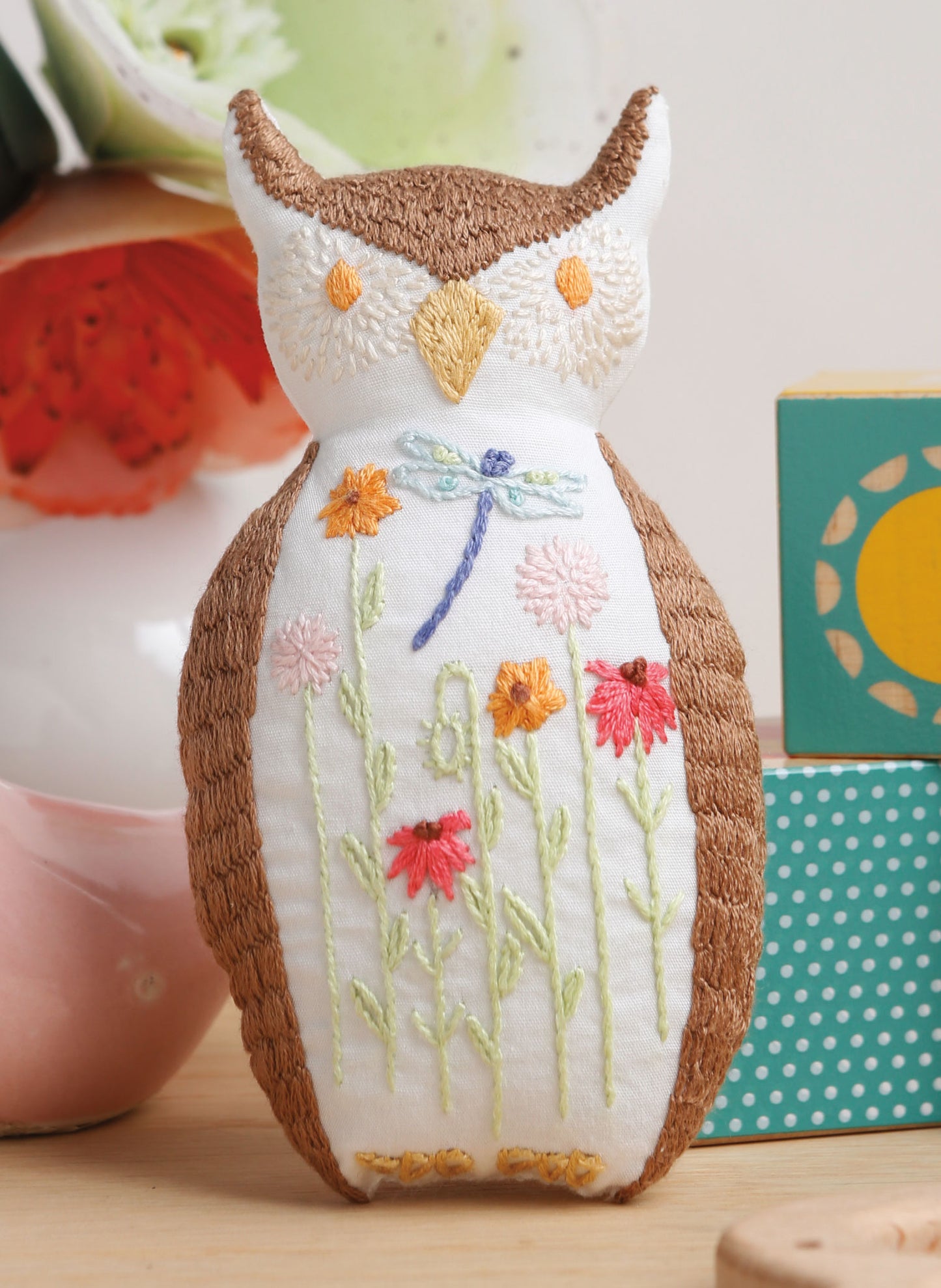 A plush owl doll with animal embroidery and detailed wings, flowers, and a dragonfly sits near blocks and a vase, inspired by the "Forest Friends—12 Adorable Animals to Embroider by Hand" digital download from Leisure Arts.