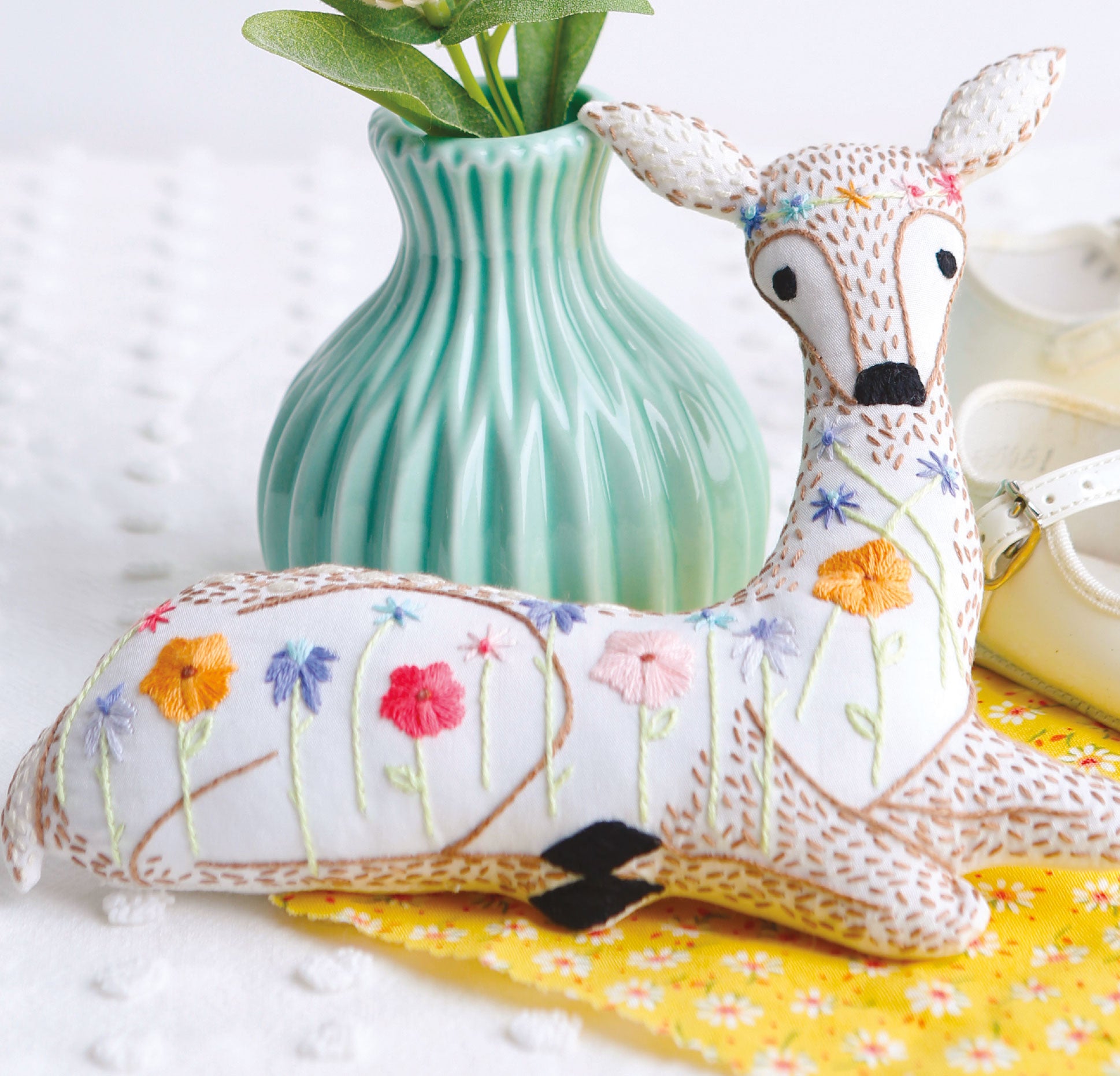 A plush deer with animal embroidery and colorful flowers, inspired by Leisure Arts' "Forest Friends—12 Adorable Animals to Embroider by Hand" digital download, sits in front of a green plant vase on a white surface with a yellow cloth.