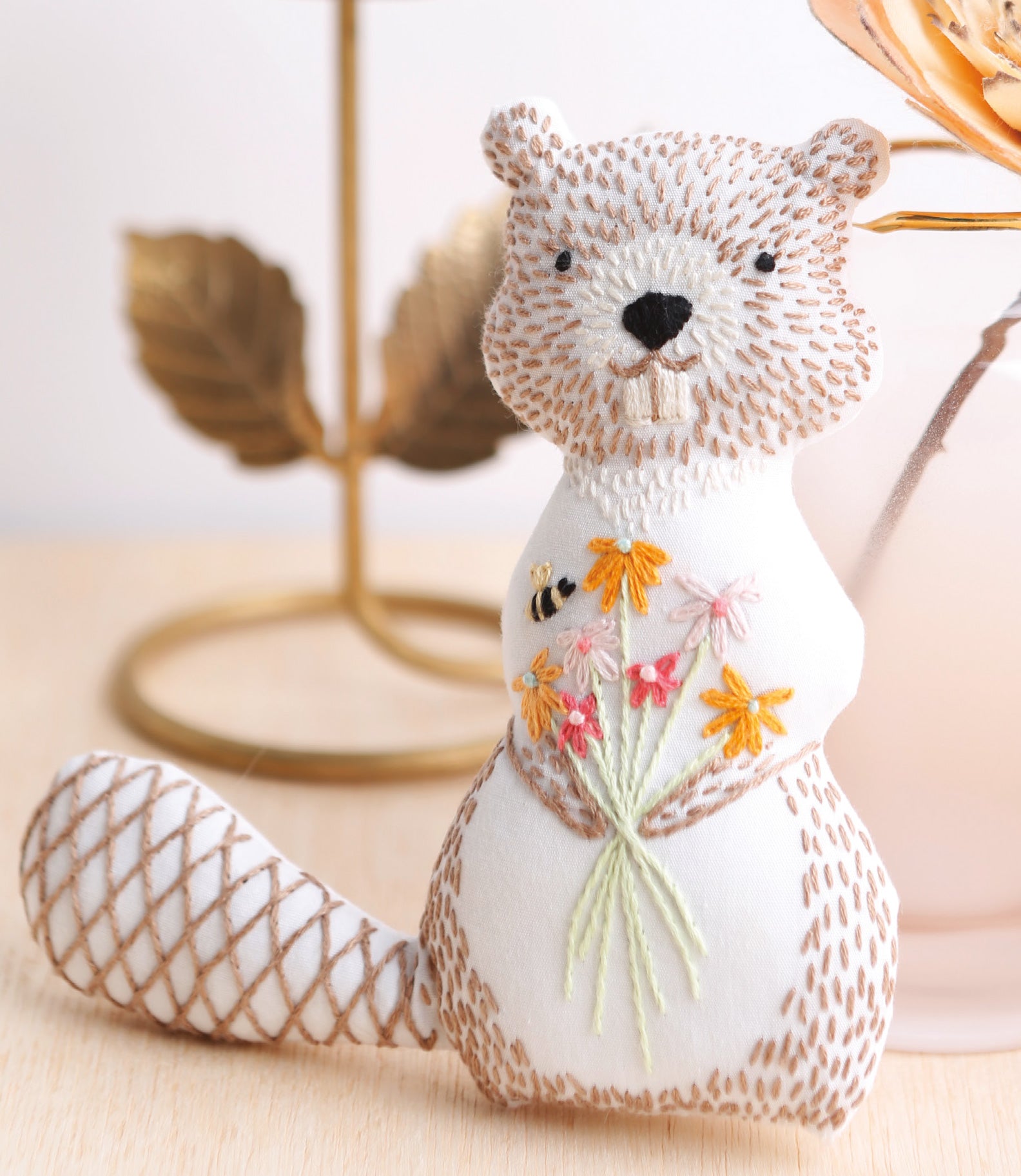 A plush beaver inspired by "Forest Friends—12 Adorable Animals to Embroider by Hand" from Leisure Arts features embroidered details, holds a flower bouquet and bee, and sits on a light wood surface with gold decor in the background.