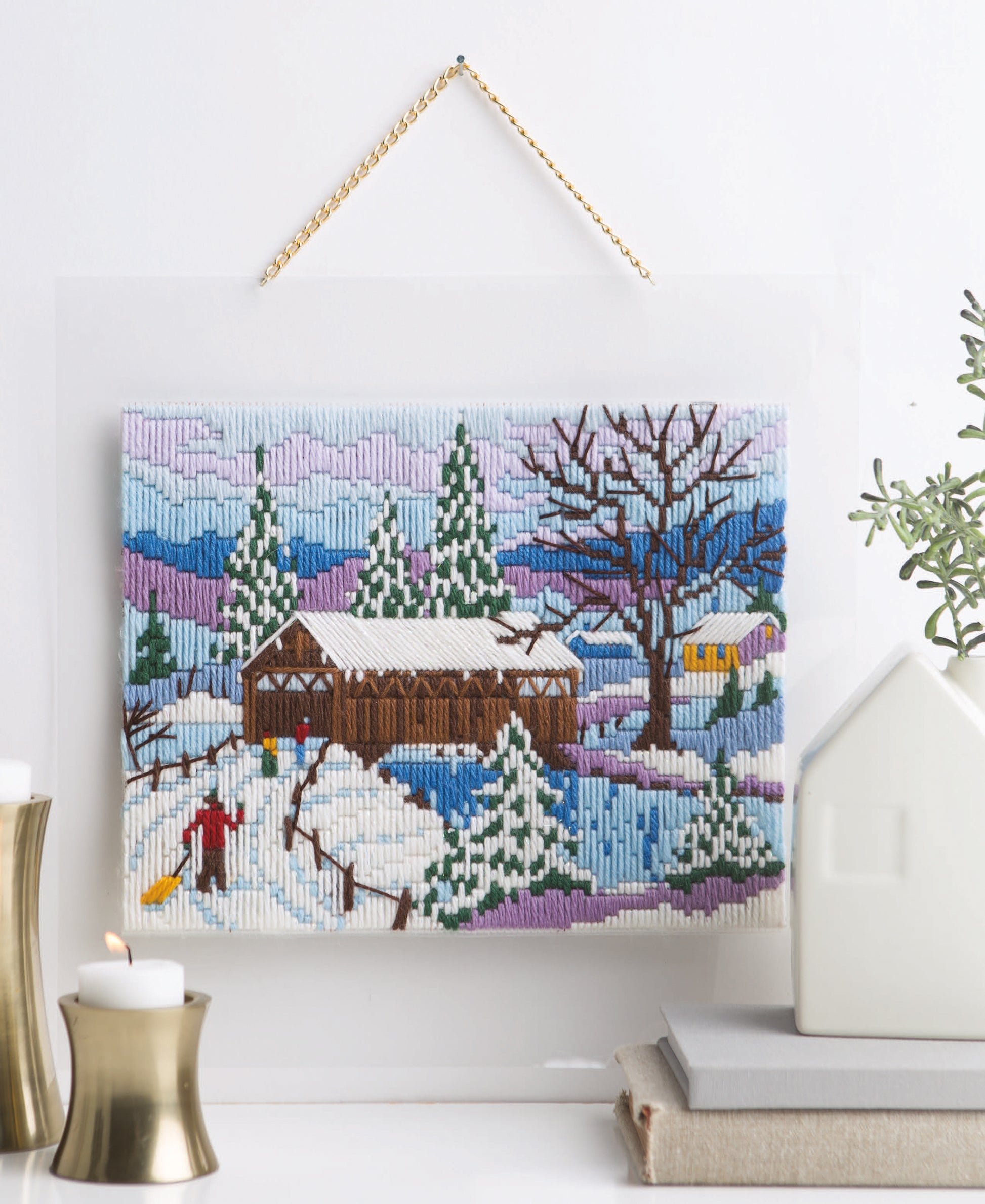 Lovely Seasons in Long-Stitch by Leisure Arts features an embroidered winter scene with snowy trees, a covered bridge, and people. Candles, books, a white house ornament, and a small plant sit on the surface below the framed artwork.