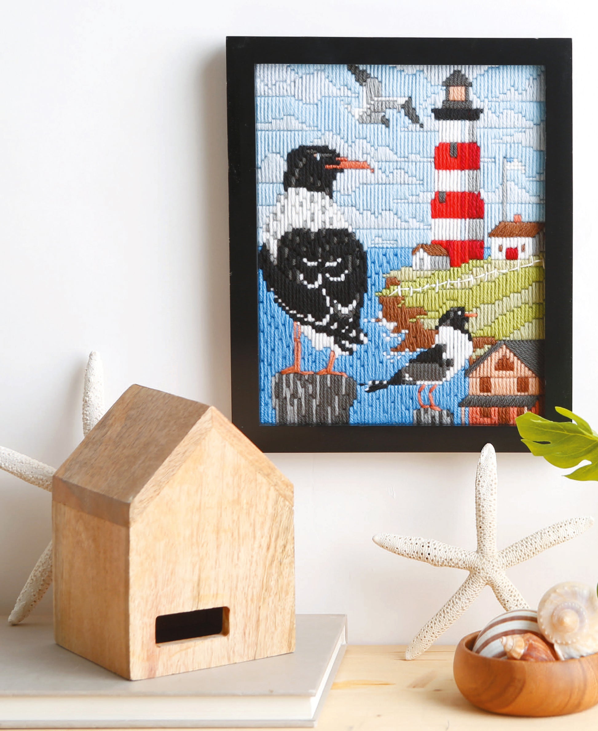 A framed Leisure Arts "Lovely Seasons in Long-Stitch" seabirds and lighthouse piece hangs above a shelf adorned with starfish, a wooden house, shells in a glass dish, and a green leaf.