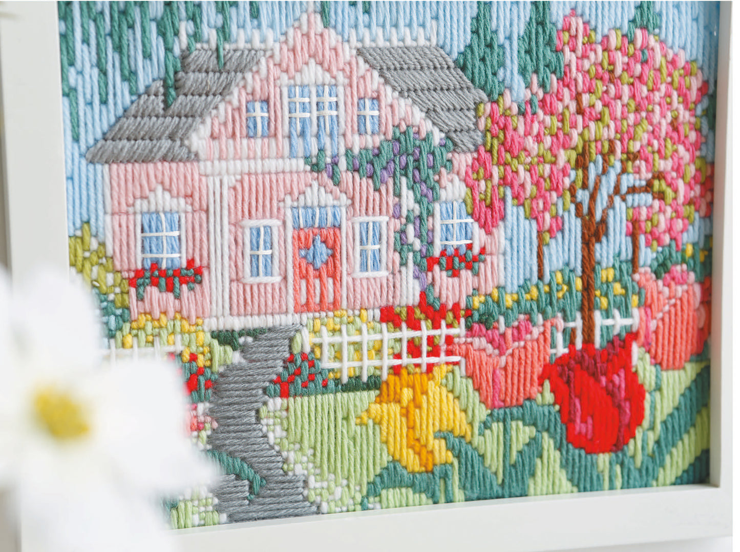 Create a charming scene of a pink house with white trim, gardens, and a picket fence under blue skies with Lovely Seasons in Long-Stitch by Leisure Arts, inspired by plastic canvas designs and featuring flowers along the path.
