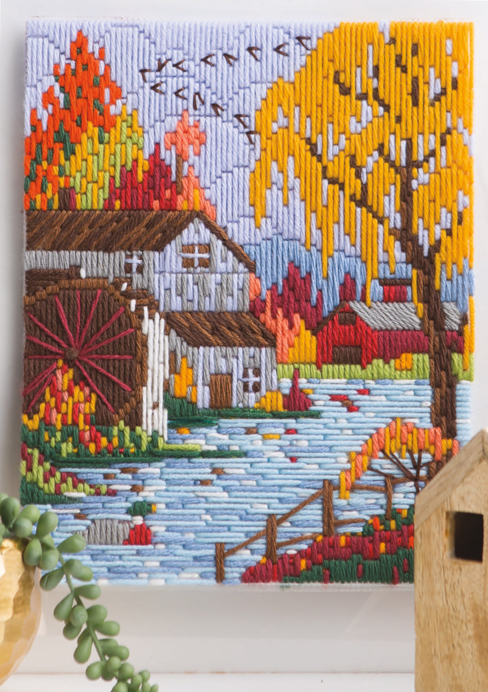Lovely Seasons in Long-Stitch by Leisure Arts features colorful embroidery of an autumn landscape with a watermill, river, vibrant trees, birds, and distant houses—capturing four seasons in charming long stitch designs.
